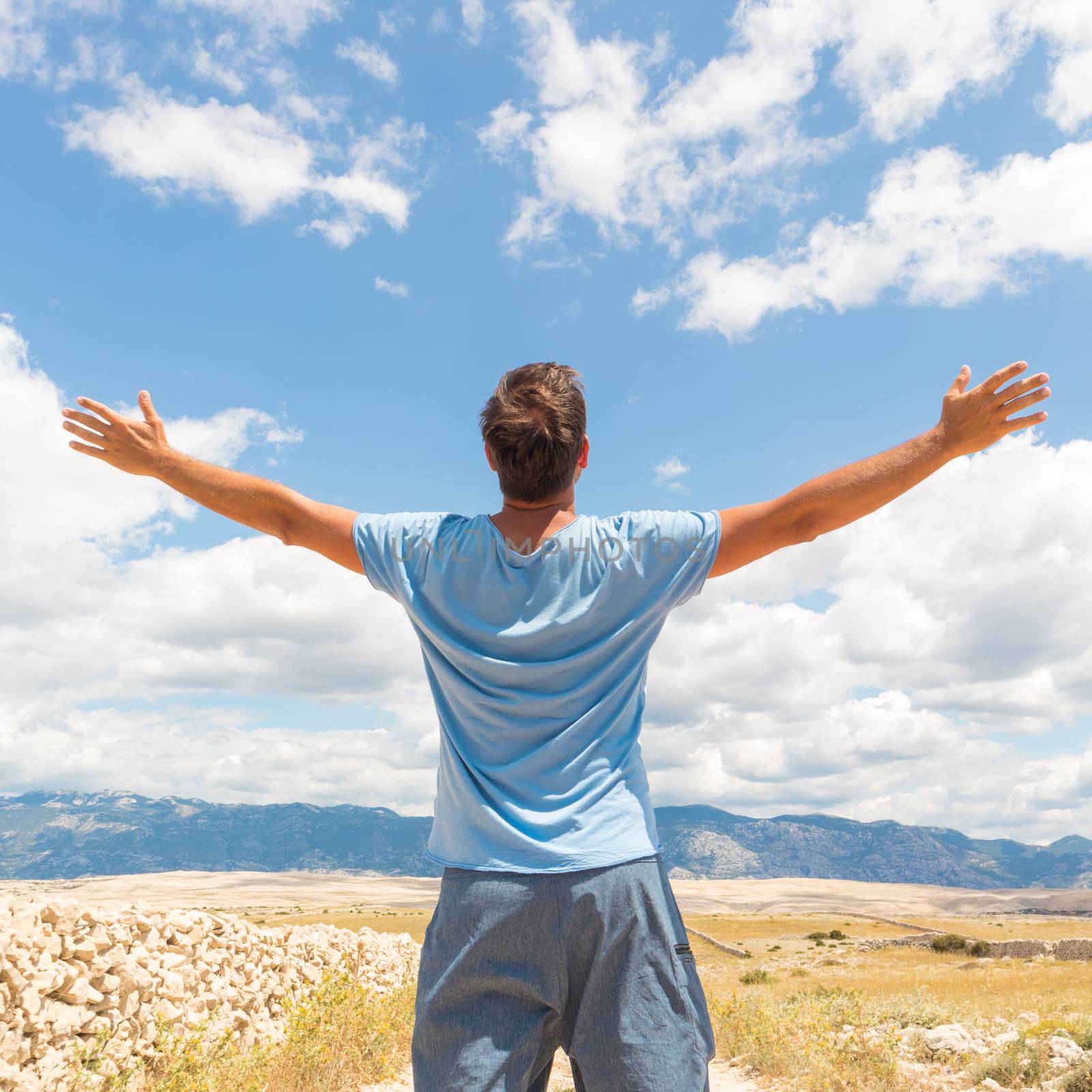 Rear view of casual sporty man standing on a dirt country road rising hands up to the clouds on a blue summer sky. Freedom and travel adventure concept. by kasto