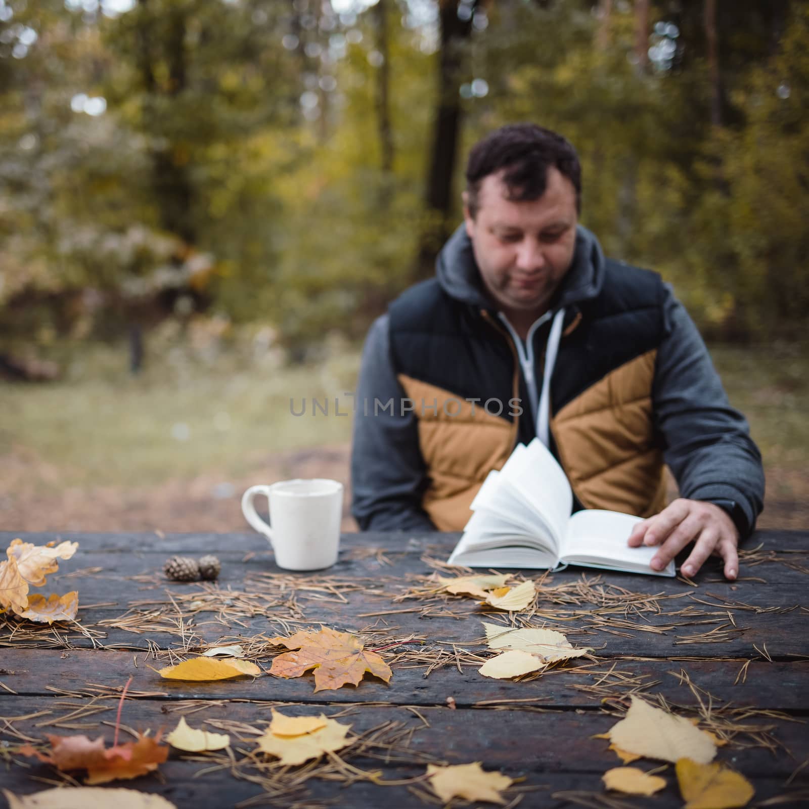 a young man sits in an autumn park at an old wooden table, drinks tea and reads a book. Warm dark autumn atmosphere, soft focus. In blur, copy space by Tanacha