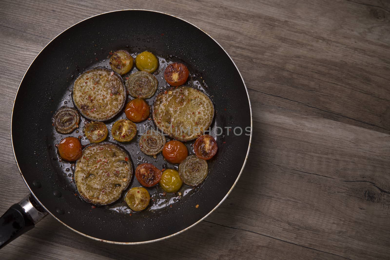Frying pan with fried aubergine cherry tomatos and onion on a wooden table, top view
