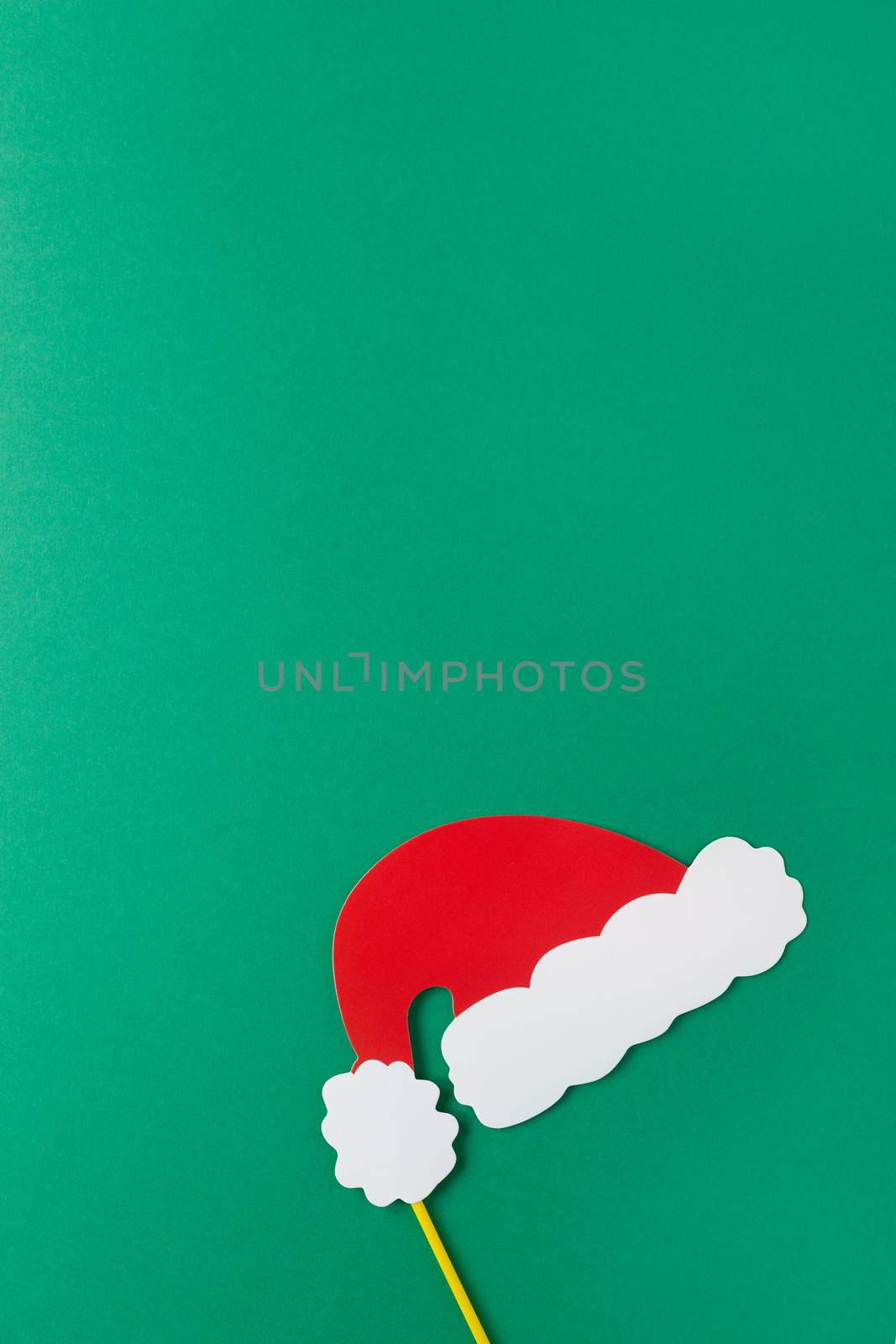 Christmas decoration, red Santas hat on stick on green background with copy space. Festive concept, party accessories. Vertical, flat lay. Minimal style. Top view.