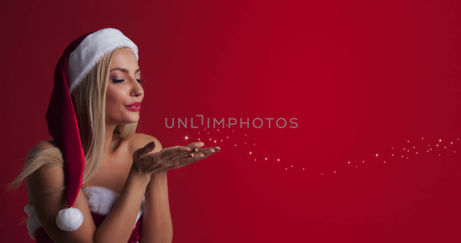 Beautiful sexy woman in santa hat and red dress blowing fairy dust off her palms over red background with copy space