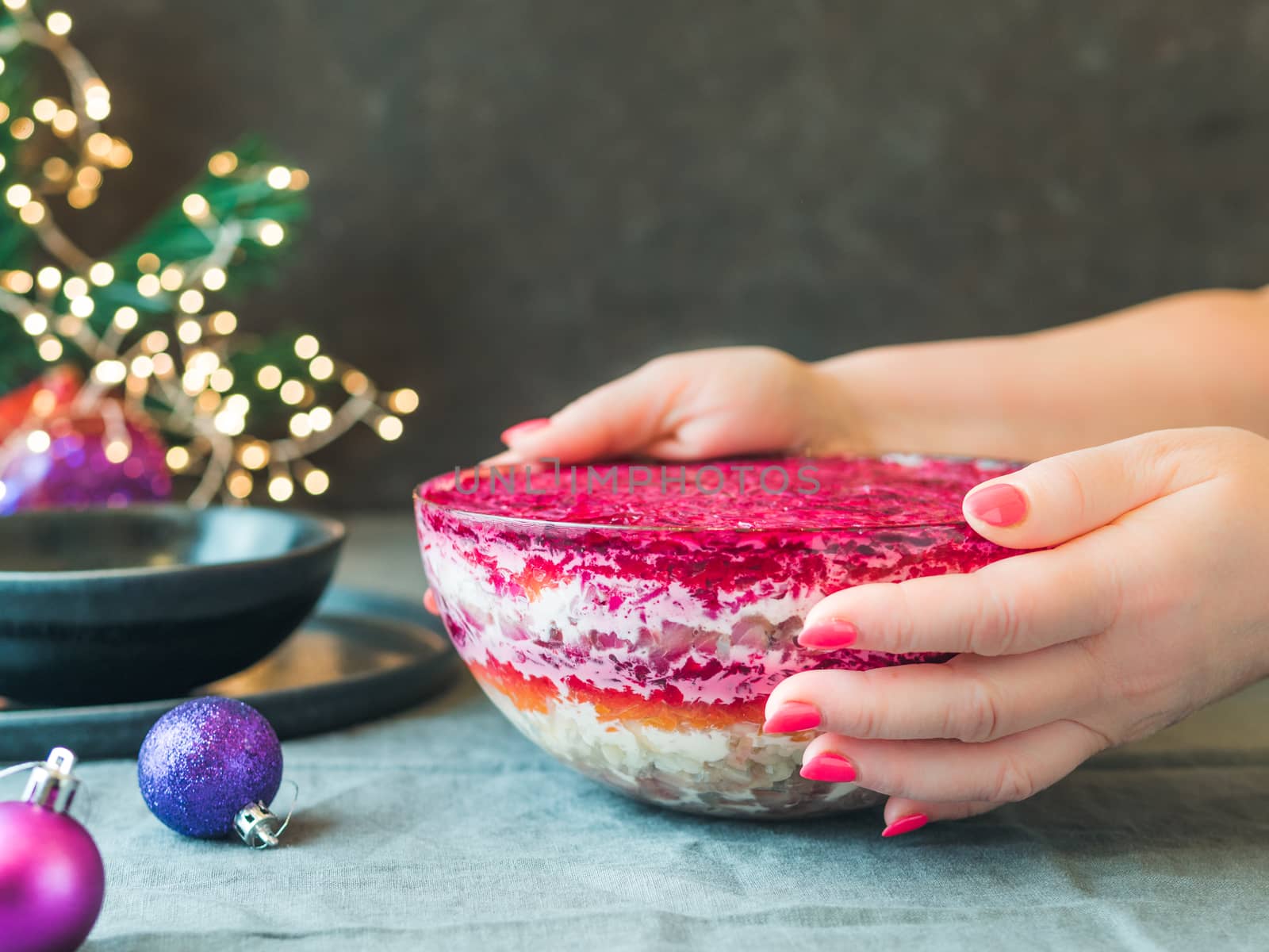 Woman hands with layered salad herring under a fur coat on festive table. Traditional russian salad with herring and vegetables in glass bowl. Copy space for text.