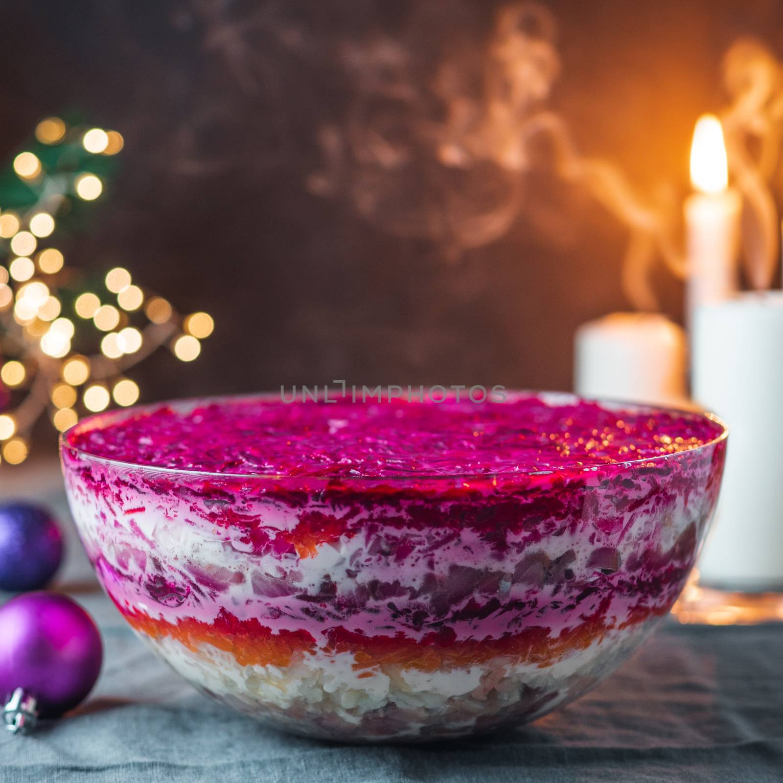 Layered salad herring under a fur coat on festive table, with candle, fir tree and light garland. Traditional russian salad with herring and vegetables in glass bowl. Copy space for text.