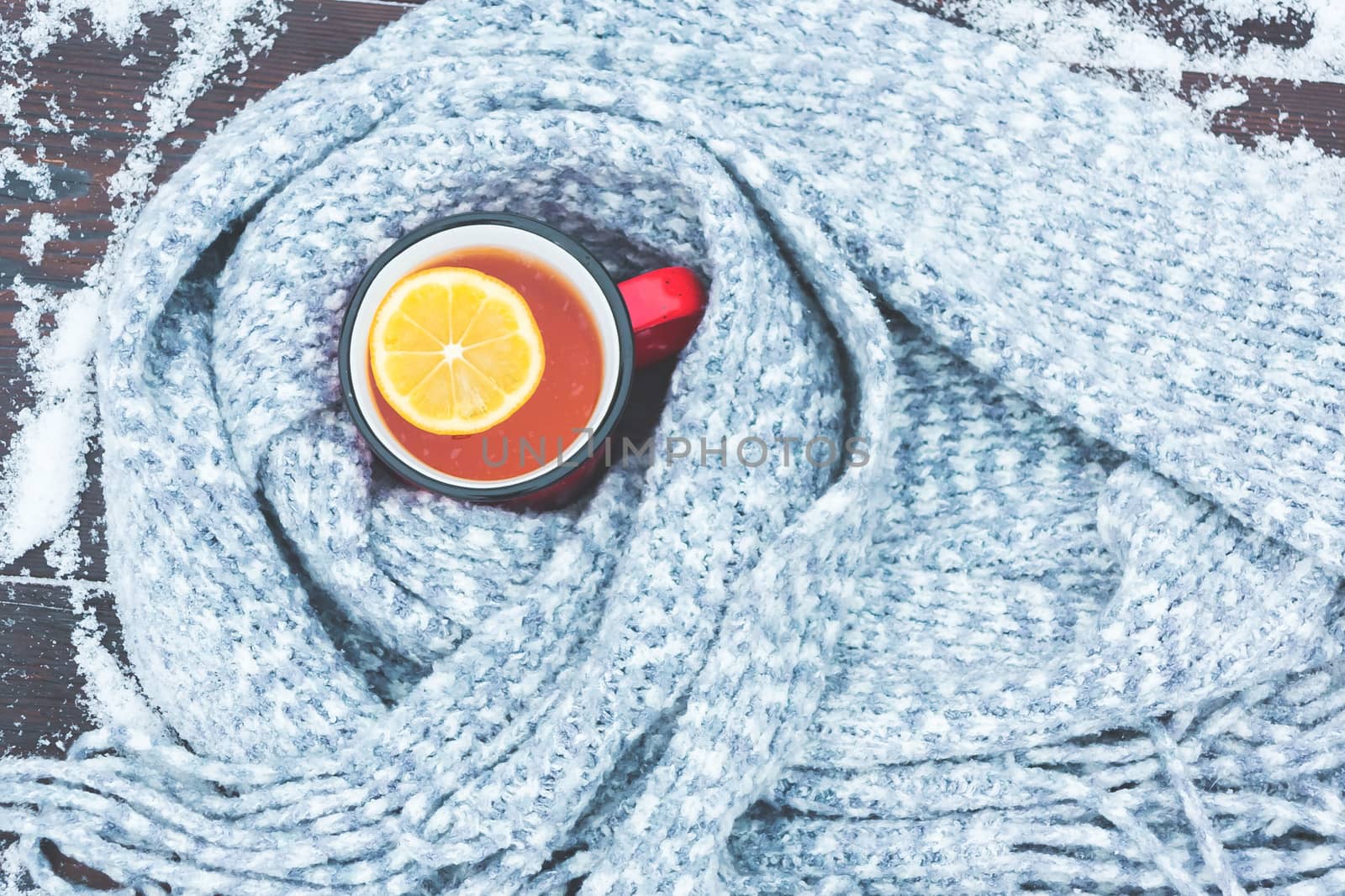 Red enameled cup of hot tea with lemon wrapped in a knitted scarf on a snowy wooden table.