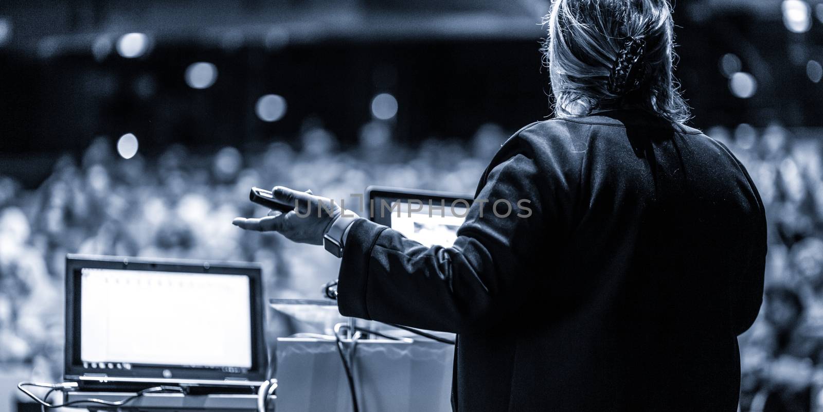 Female speaker giving a talk on corporate business conference. Unrecognizable people in audience at conference hall. Business and Entrepreneurship event. Black and white, blue toned image.