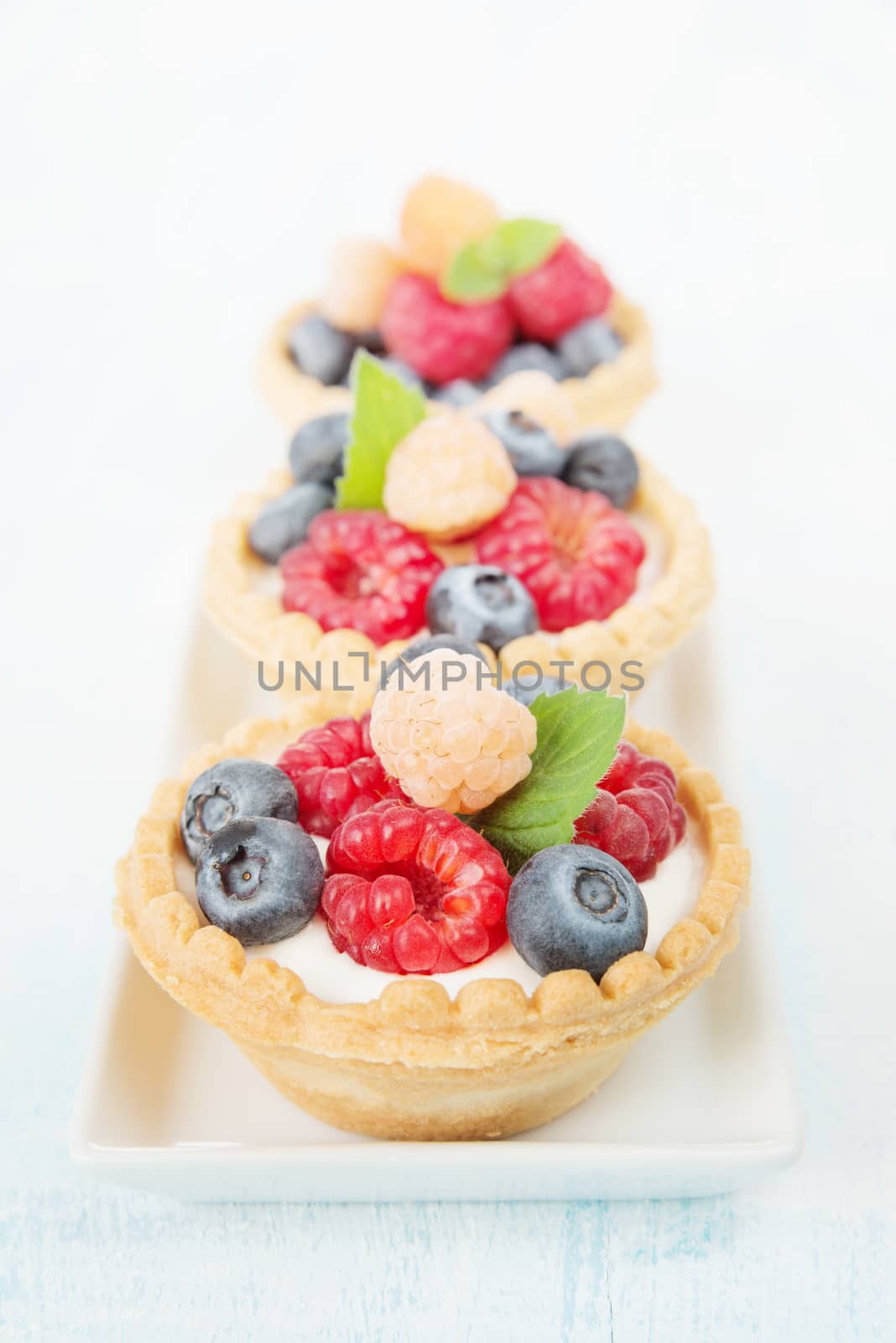 Three fruit tartlets with cream, red and yellow raspberries and blueberries in a row on a white porcelain rectangular plate closeup