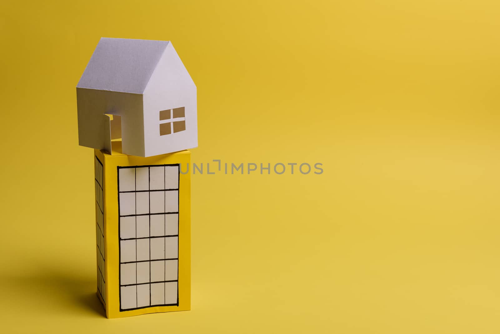 White family paper house over block of flats on yellow background paper. Minimalistic and simple concept, style. Copy space. Horizontal orientation.