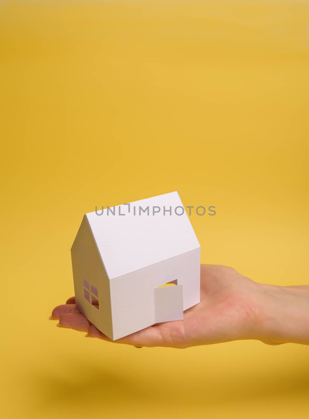 White family paper house in man hand on yellow background paper. Minimalistic style. Copy space. View from above. Vertical orientation