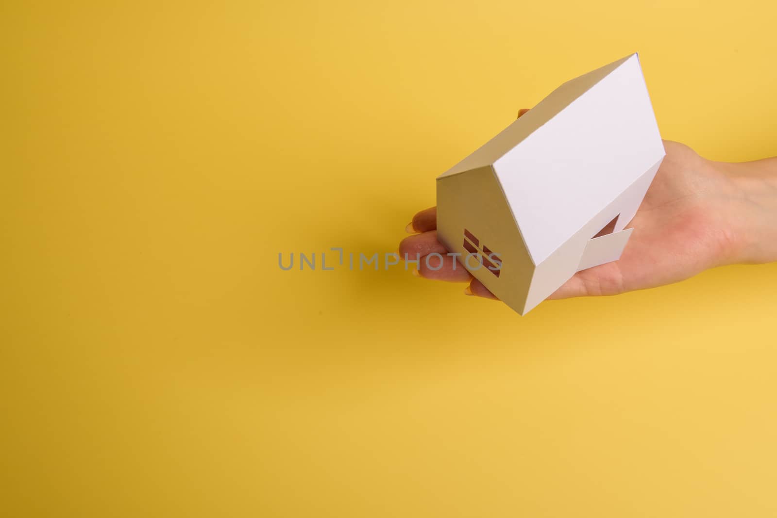 White family paper house in man hand on yellow background paper. Minimalistic style. Copy space. View from above. Horizontal orientation
