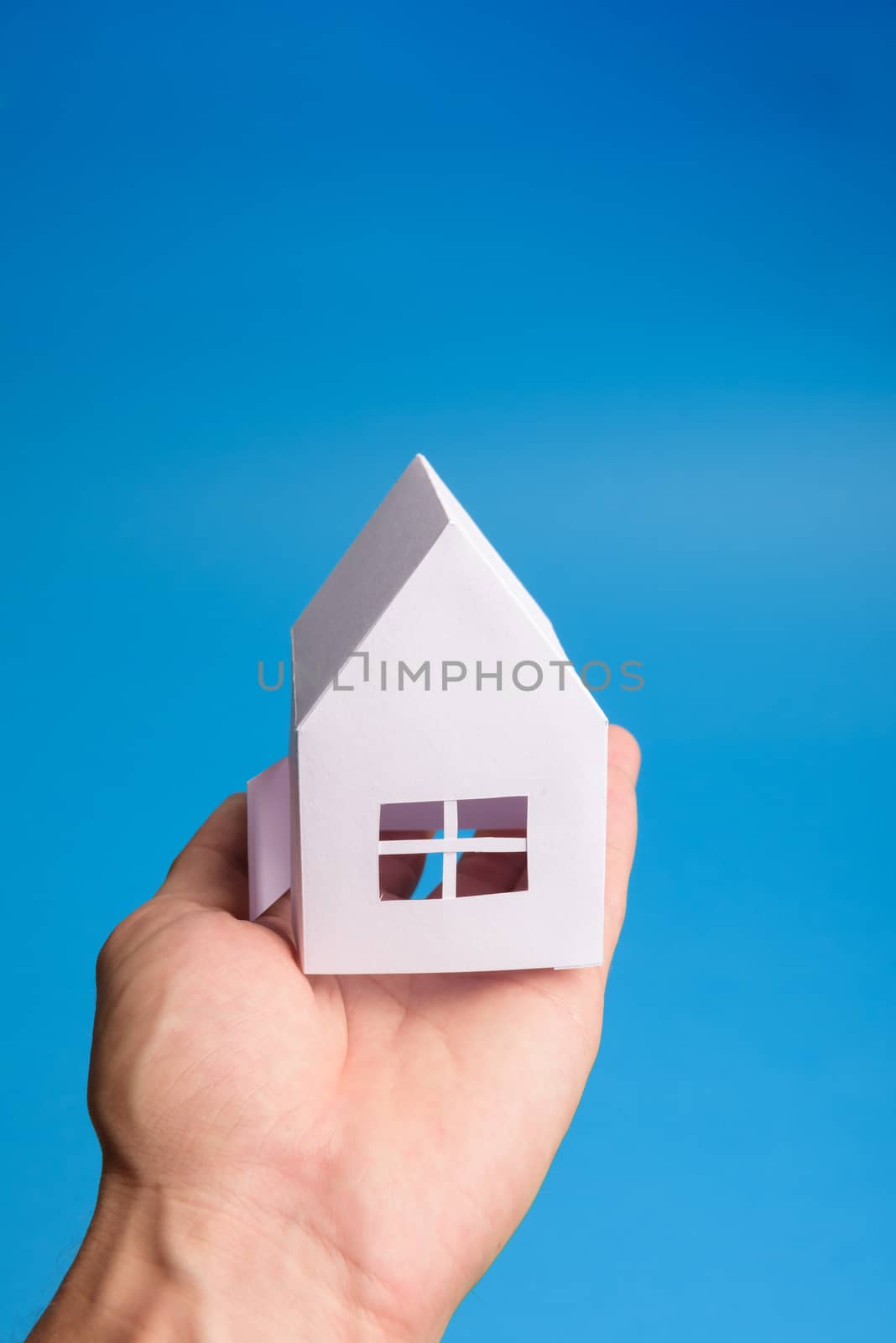 White family paper house in man hand on blue background paper. Minimalistic style. Copy space. View from above. Vertical orientation
