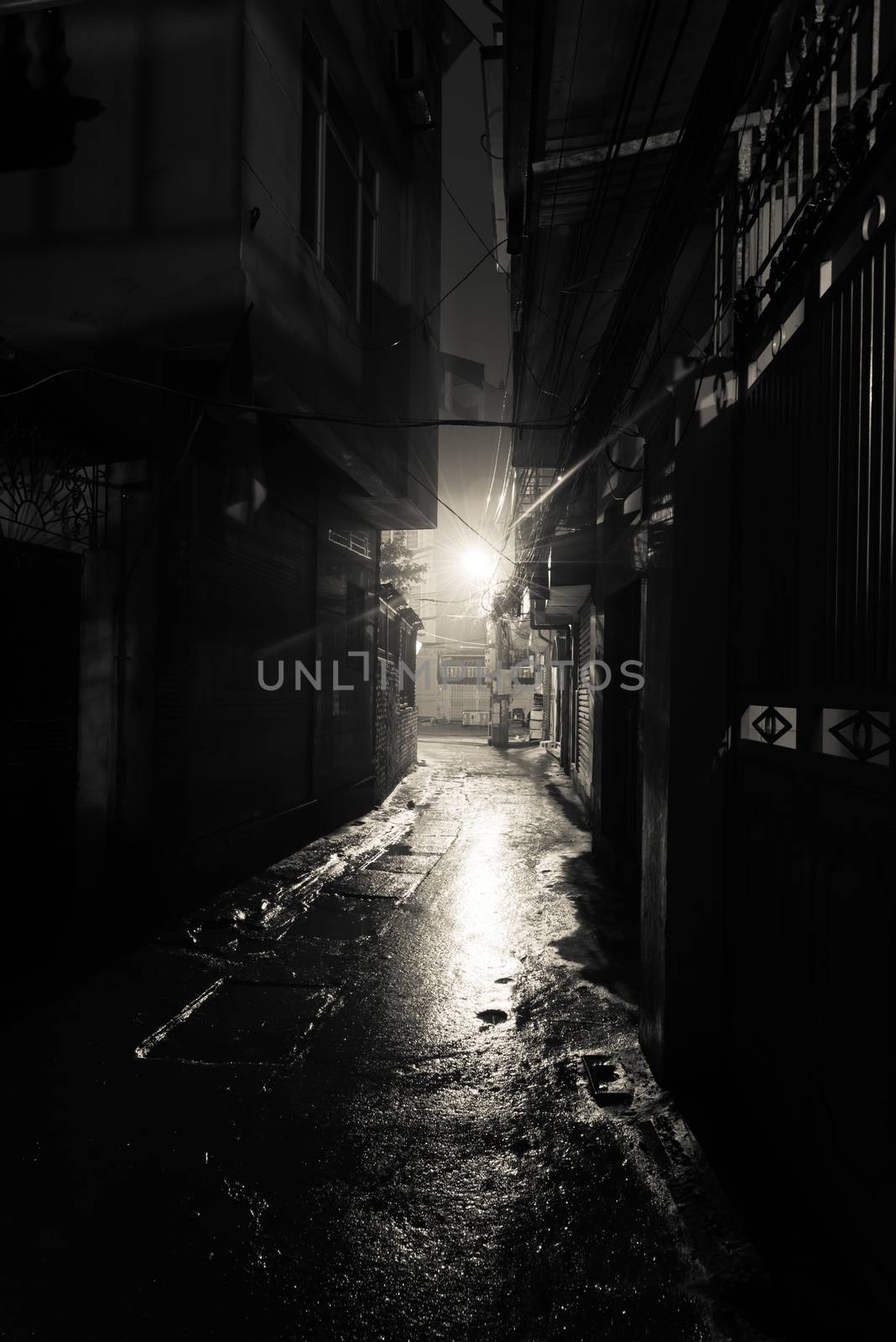 A dark, shadowy and dangerous looking urban back-alley at night time in suburbs Hanoi, Vietnam. Low light reflected on wet pavement from post lamp at the end of long road corner