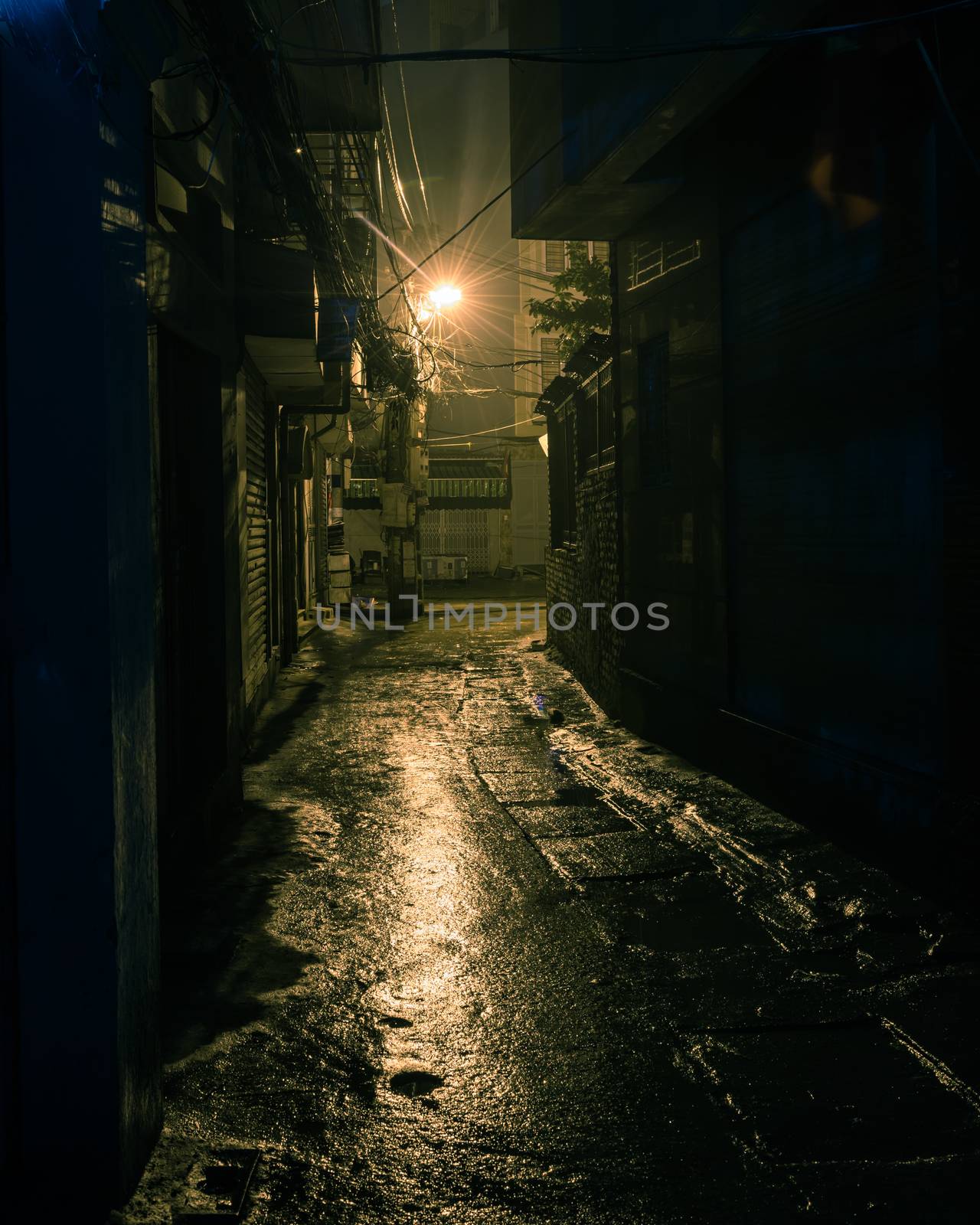 Vintage tone a dark, shadowy and dangerous looking urban back-alley at night time in suburbs Hanoi, Vietnam. Low light reflected on wet pavement from post lamp at the end of long road corner