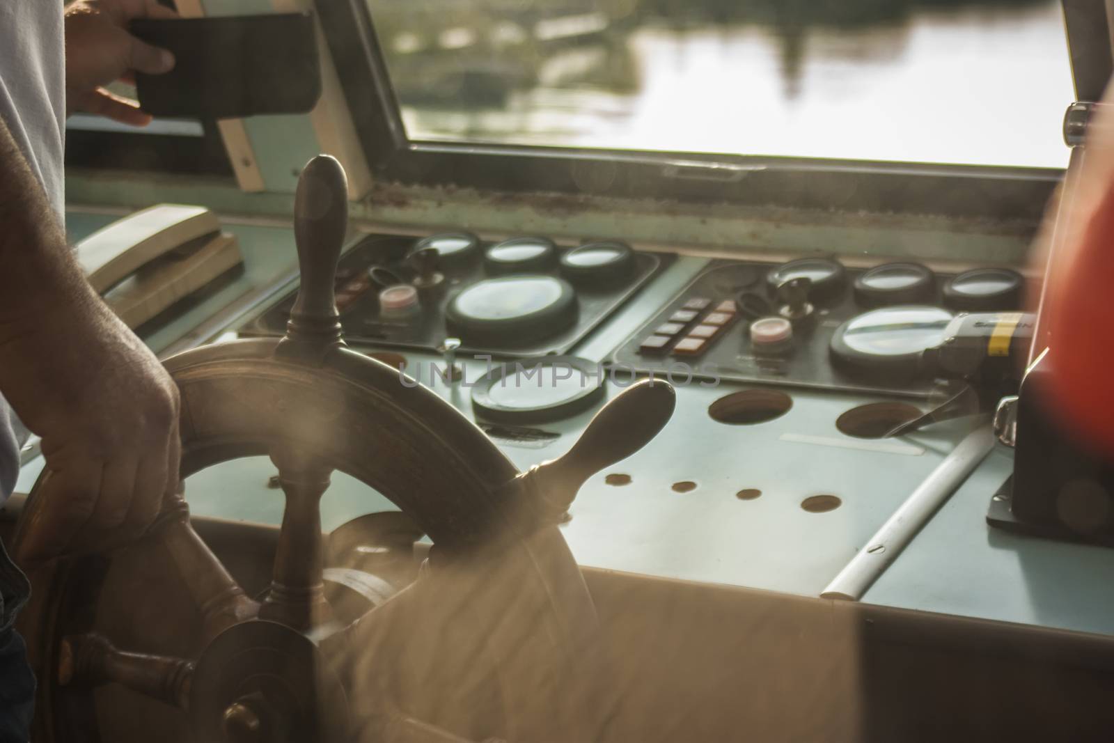 Helm of a ship in the foreground while the commander is leading the ship in a ferry on a river.