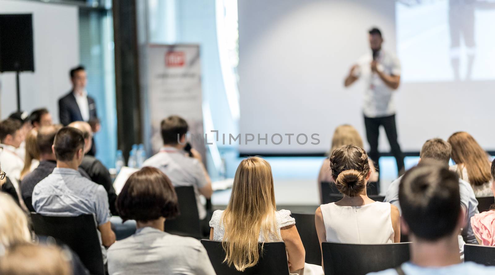 Business and entrepreneurship symposium. Speaker giving a talk at business meeting. Audience in conference hall. Rear view of unrecognized participant in audience.