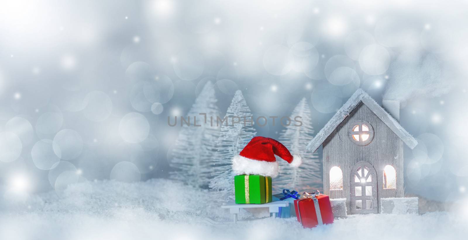 Christmas card with cozy small decorative house , gifts and Santa Claus cap on sleigh on snow over fairy tale winter fir forest background and falling snow . New Year design postcard . Copy space for text