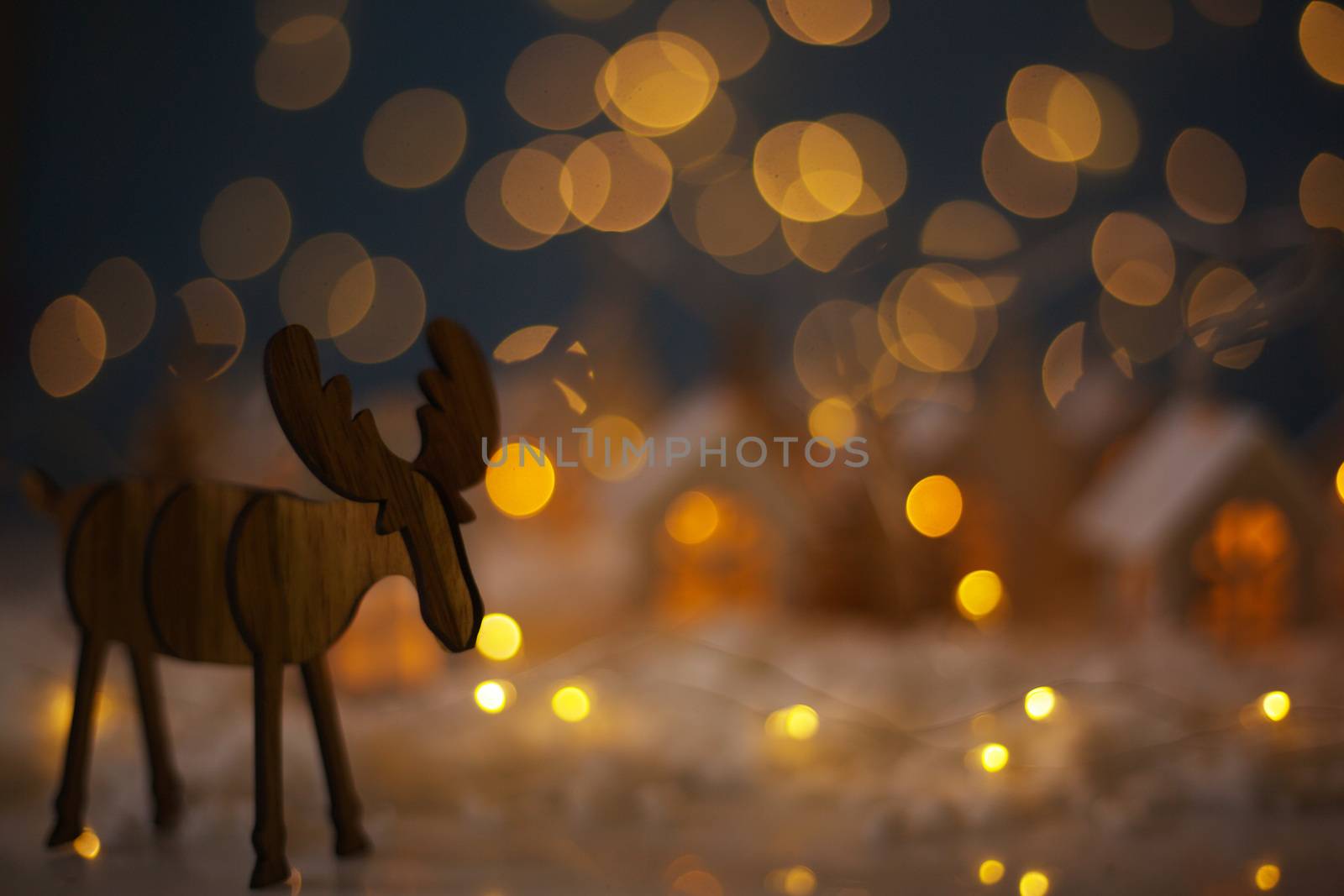 Christmas wooden reindeer over small glowing toy houses village garland over bokeh lights background with copy space for text