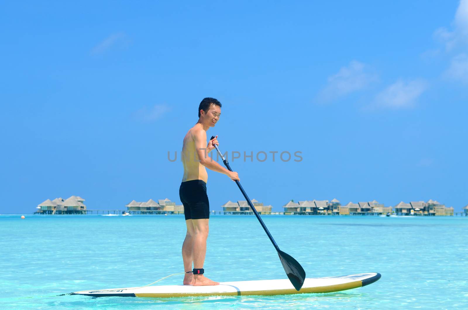 Young asian man paddle board rowing on sunny tropical paradise island with blue  sea water ocean in Maldives island.