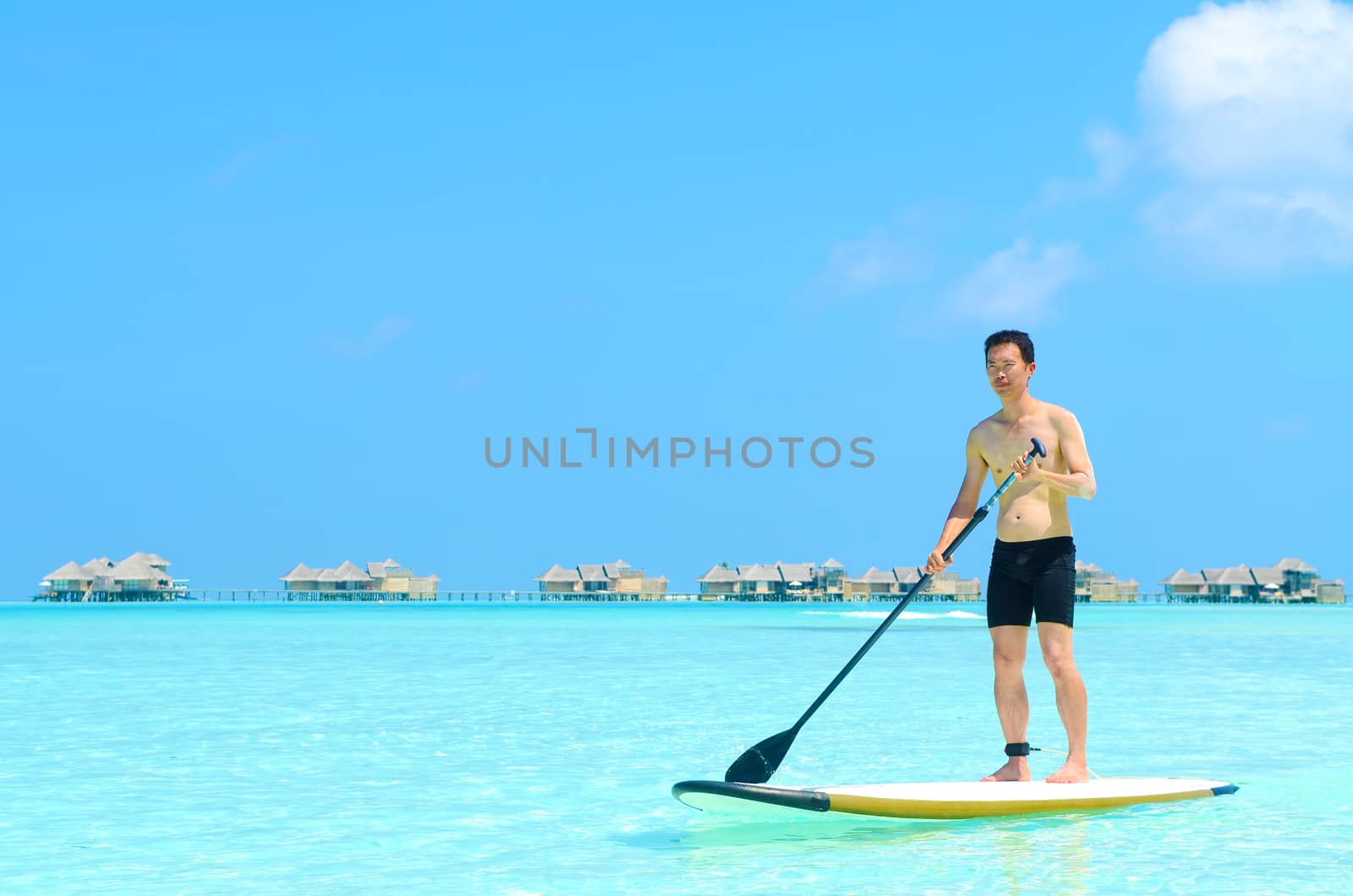 Young asian man paddle board rowing on sunny tropical paradise island with blue  sea water ocean in Maldives island.