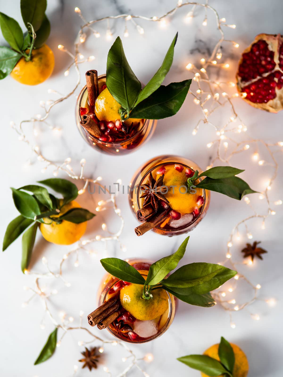 Winter sangria on white marble background. Holiday sangria in glasses with fruit slice, pomegranate and spices on decoration lighting chain tabletop. Top view or flat lay. Vertical.