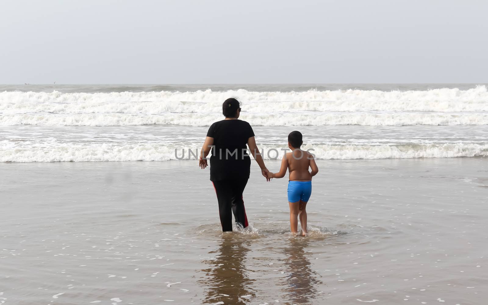 Rear View of Mother and son standing on a tropical beach in evening during sunset. The child admires his mom,s faith. Happy Mother’s day background concept. Goa, India, South Asia Pacific In Summer.