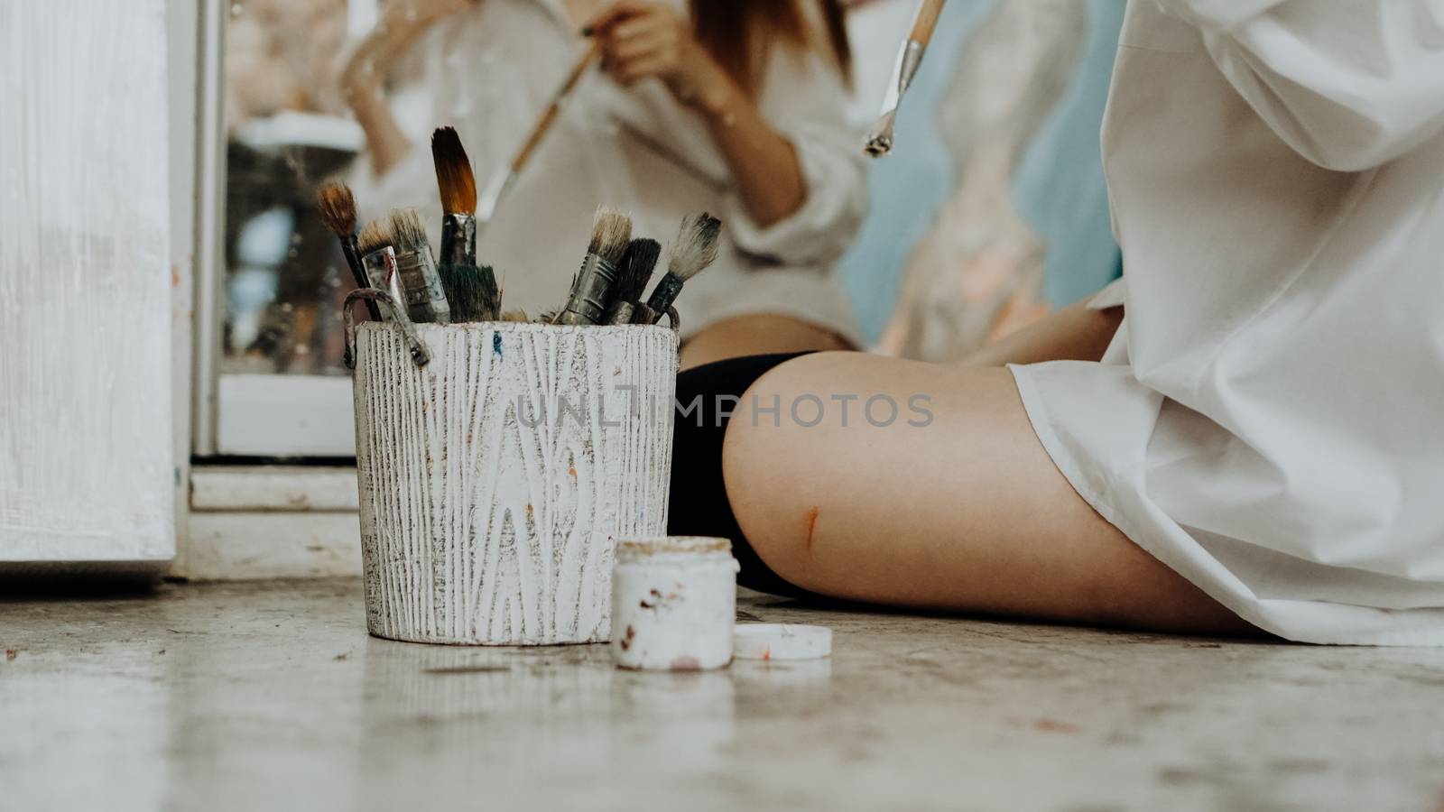 Woman painter sitting on the floor in front of mirror and drawing. Art studio interior. Drawing supplies, oil paints, artist brushes