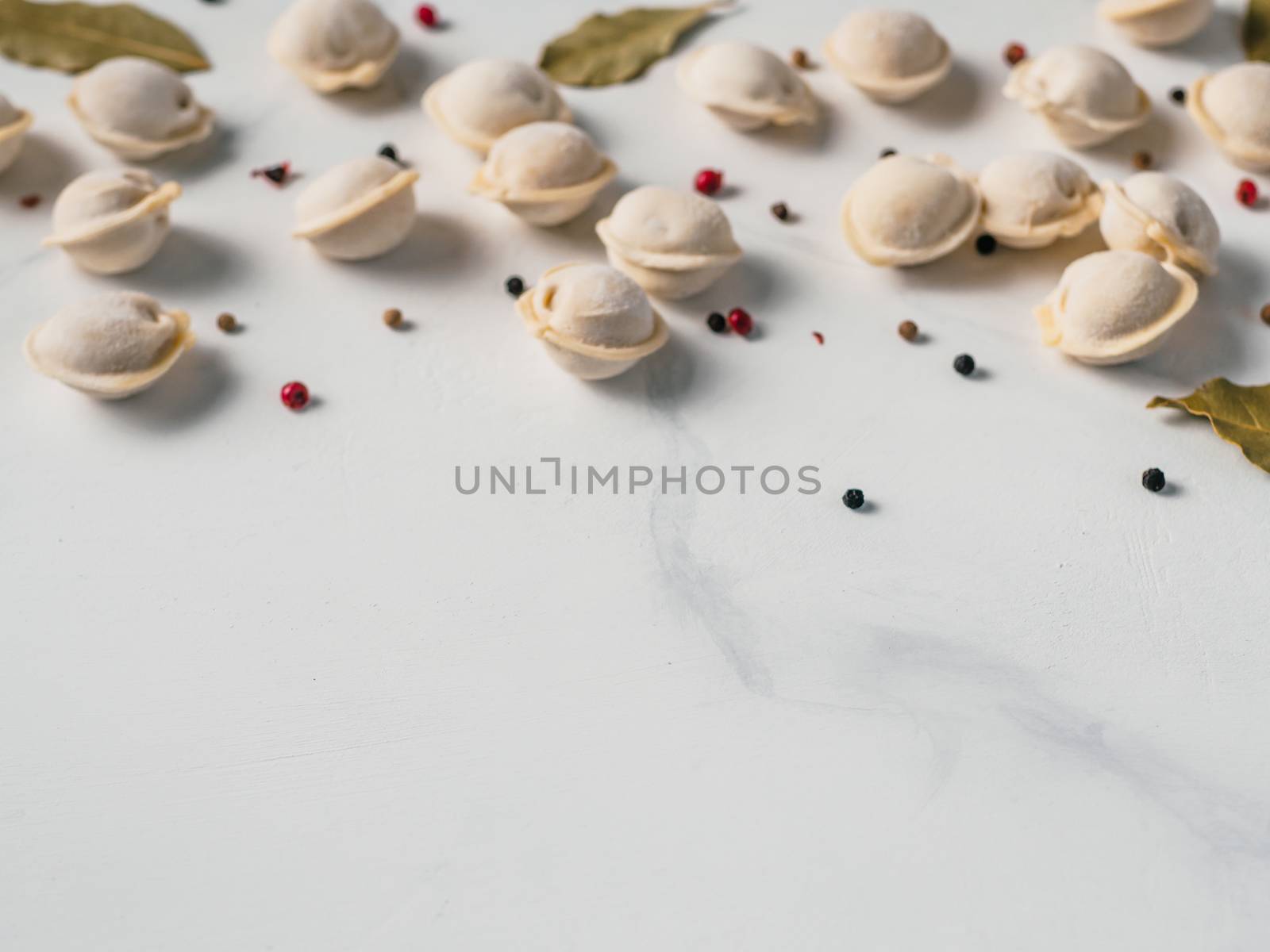 Pattern of frozen uncooked russian pelmeni with peppercorns and bay leaves on white marble table. Creative layout of dumplings. Beautiful scattered raw dumplings. Top view or flat lay. Copy space