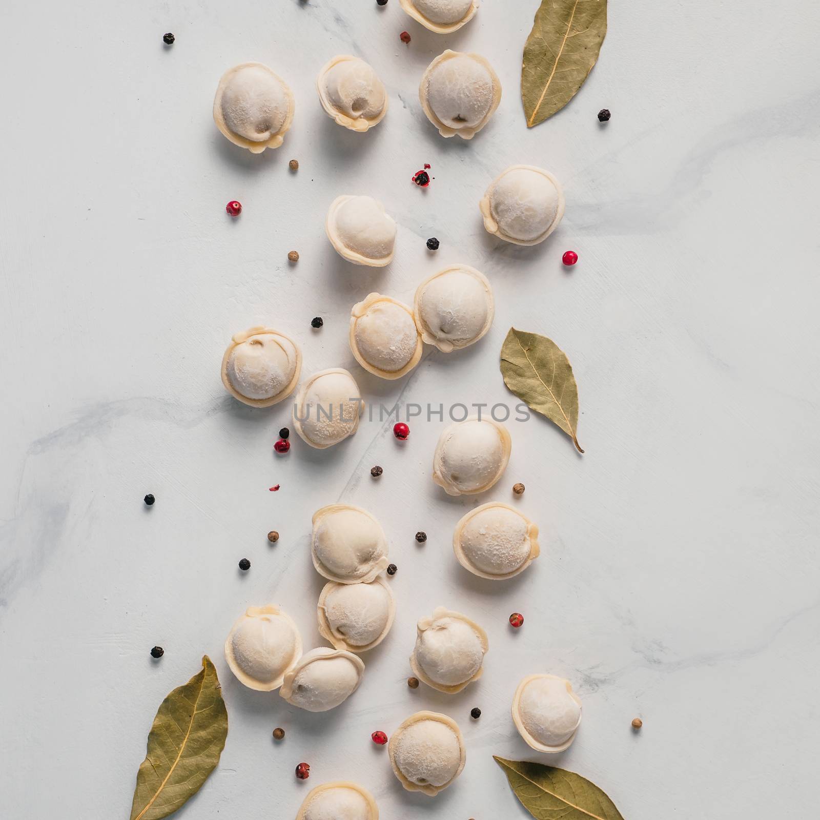 Pattern of frozen uncooked russian pelmeni with peppercorns and bay leaves on white marble table. Creative layout of dumplings. Beautiful scattered raw dumplings
