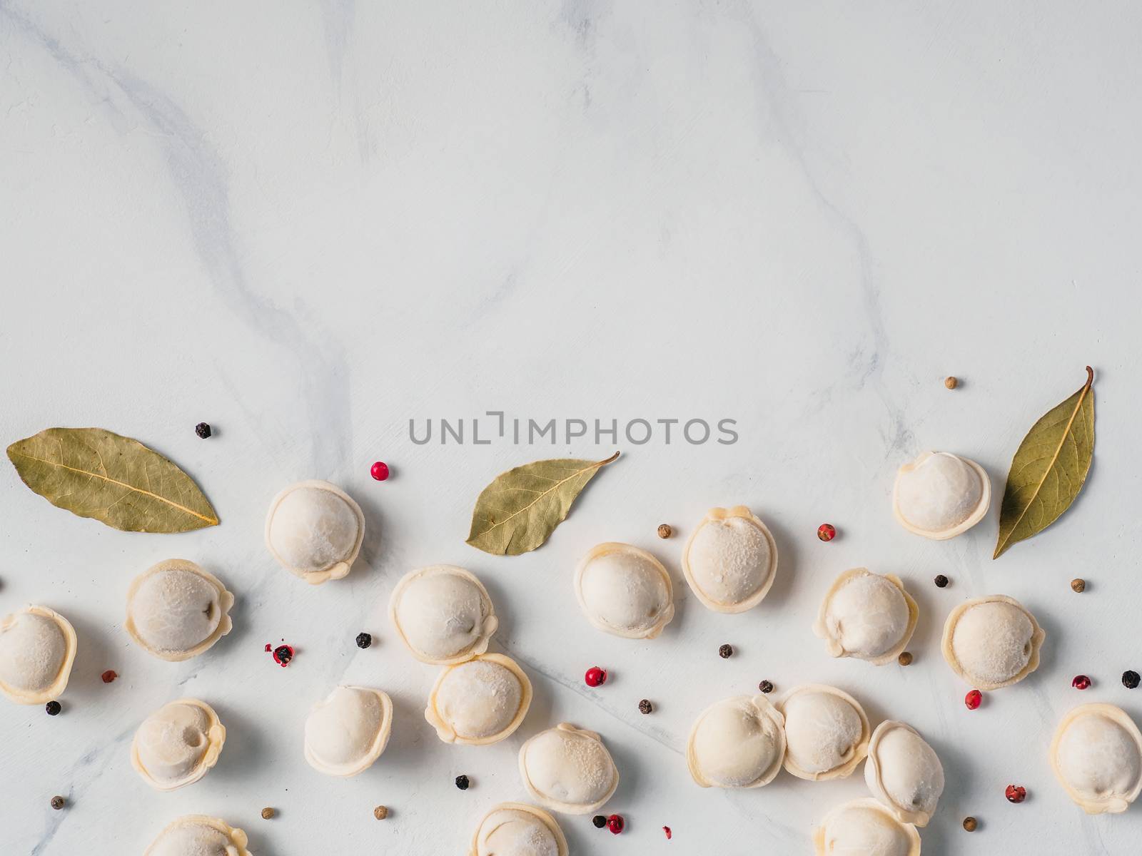 Pattern of frozen uncooked russian pelmeni with peppercorns and bay leaves on white marble table. Creative layout of dumplings. Beautiful scattered raw dumplings. Top view or flat lay. Copy space