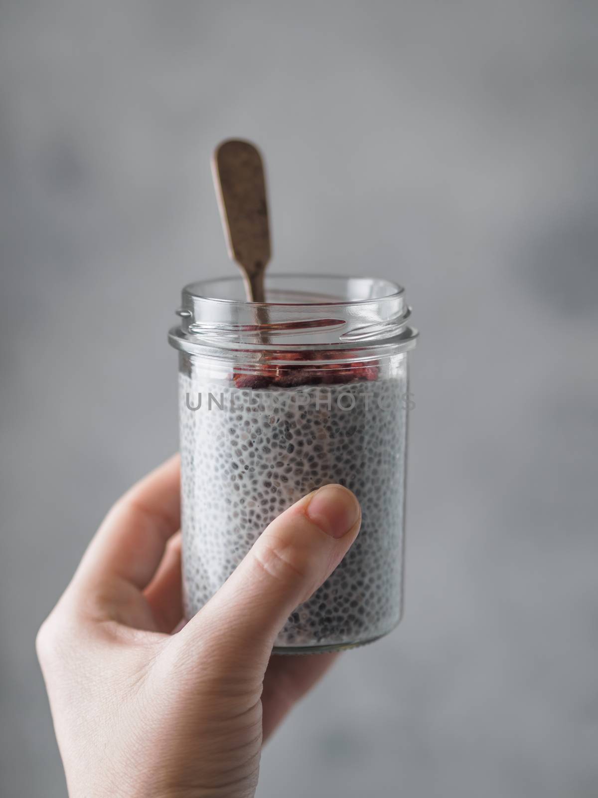 Chia pudding in woman hand on gray background. Female hand hold glass jar with chia seeds puding served goji berries. Copy space for text. Vertical.