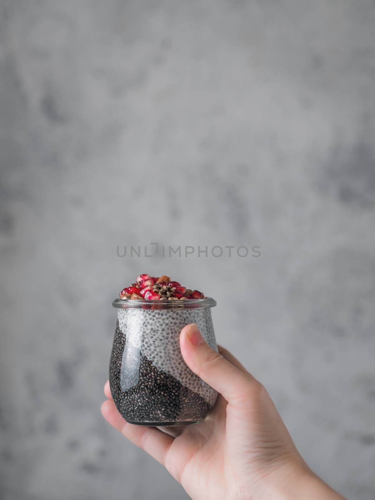 Chia pudding with black charcoal and white vanilla plant-based milk in woman hand on gray background. Female hand hold glass jar with chia puding served pomegranate and hemp. Copy space. Vertical