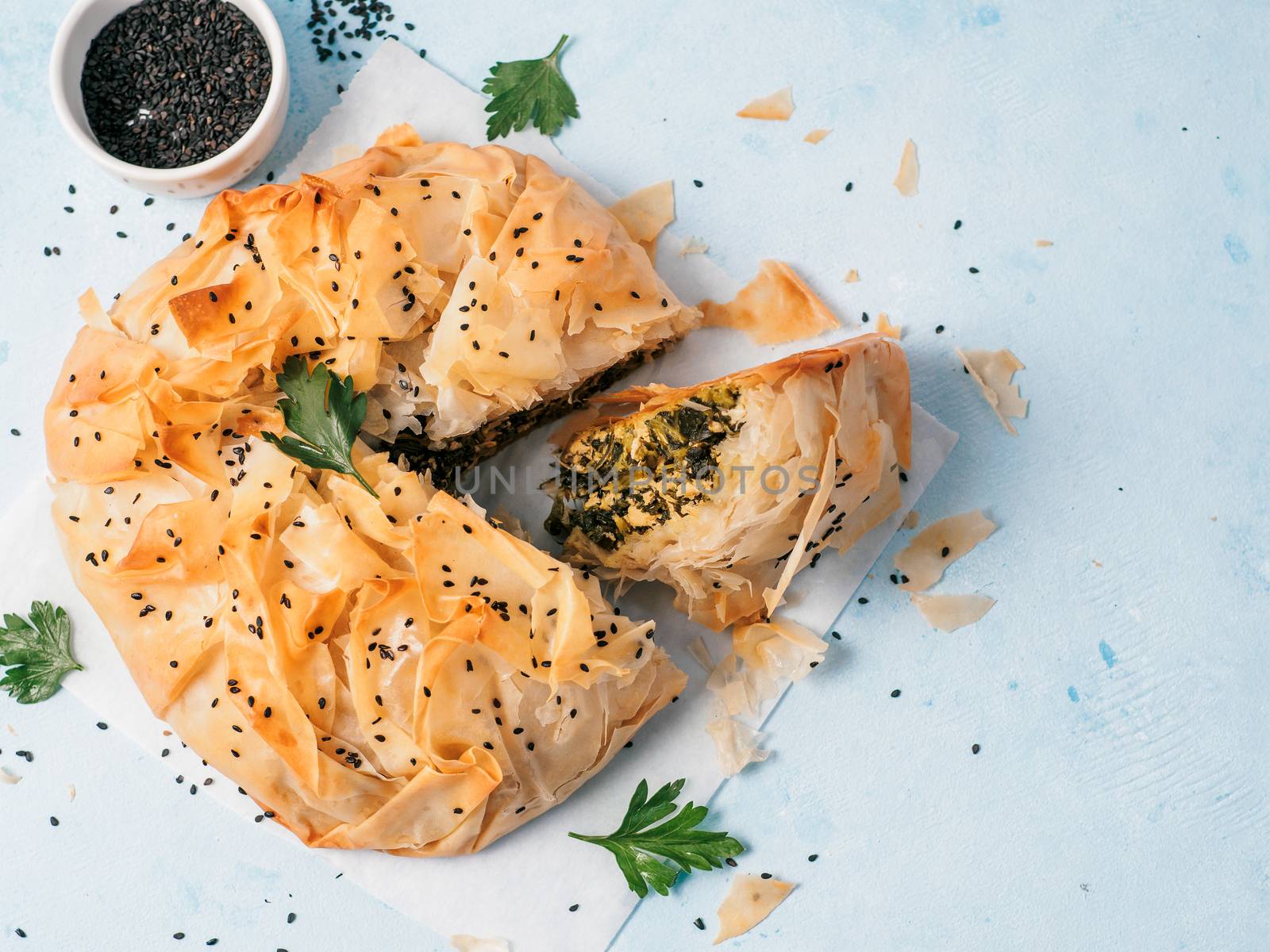 Greek pie spanakopita on white baking paper over blue background. Ideas and recipes for vegetarian or vegan Spanakopita Spinach Pie from fillo pastry cut in slices. Copy space. Top view or flat lay.