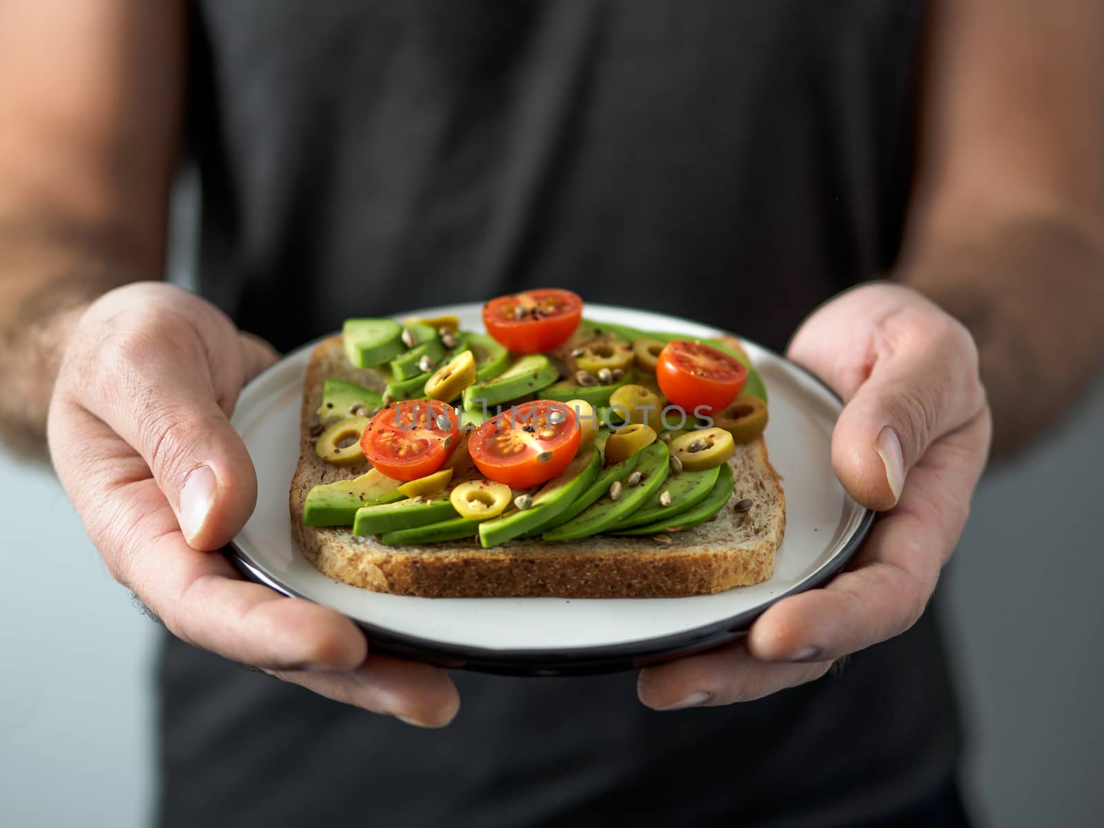 Hands takes plate with vegan sandwich. Healthy appetiezer - whole wheat bread toast with avocado, cherry tomatoes and olives