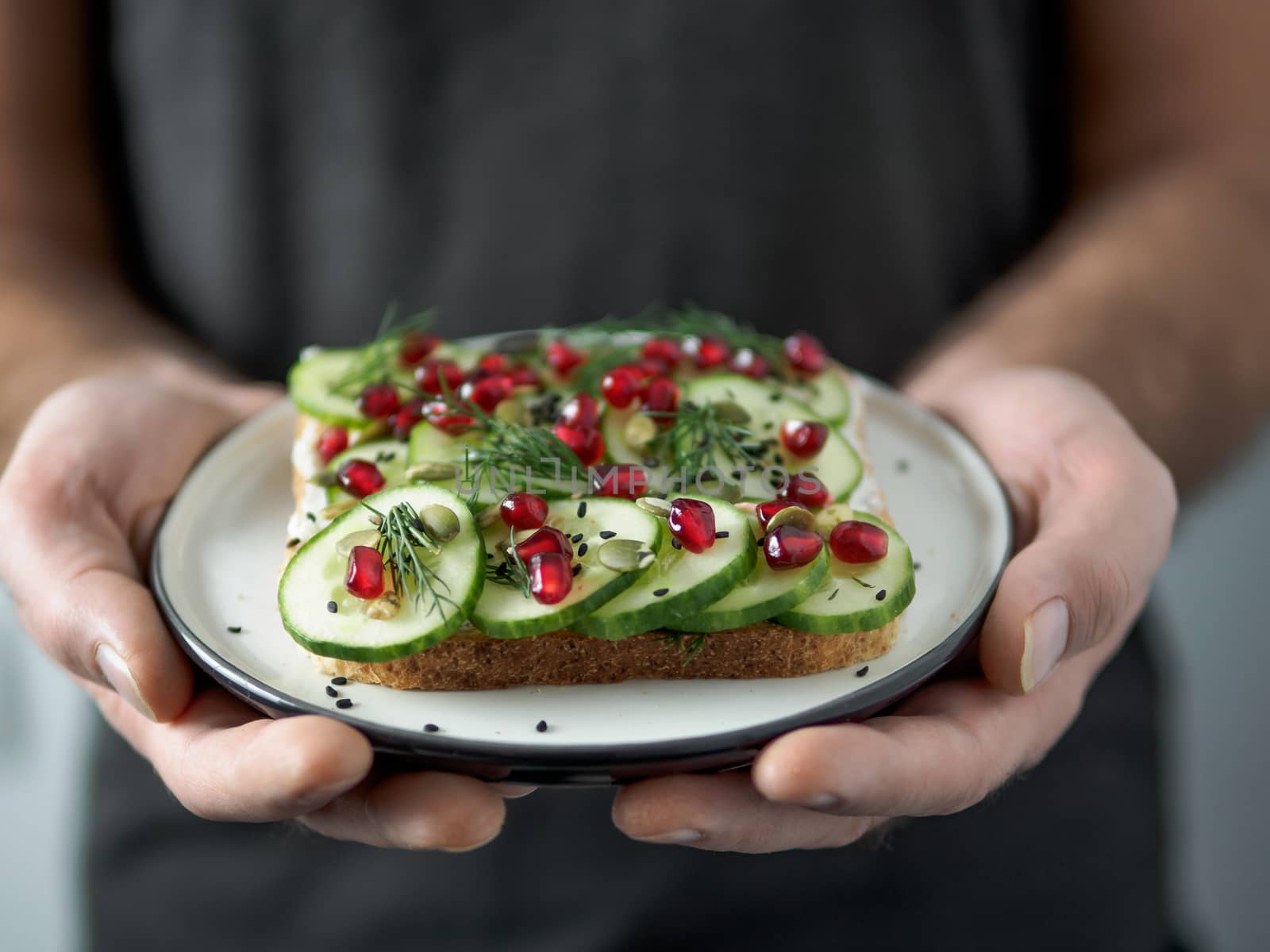 Hands takes plate with vegan sandwich. Healthy appetiezer - whole wheat bread toast with cucumber, pomegranate and black sesame. Vertical
