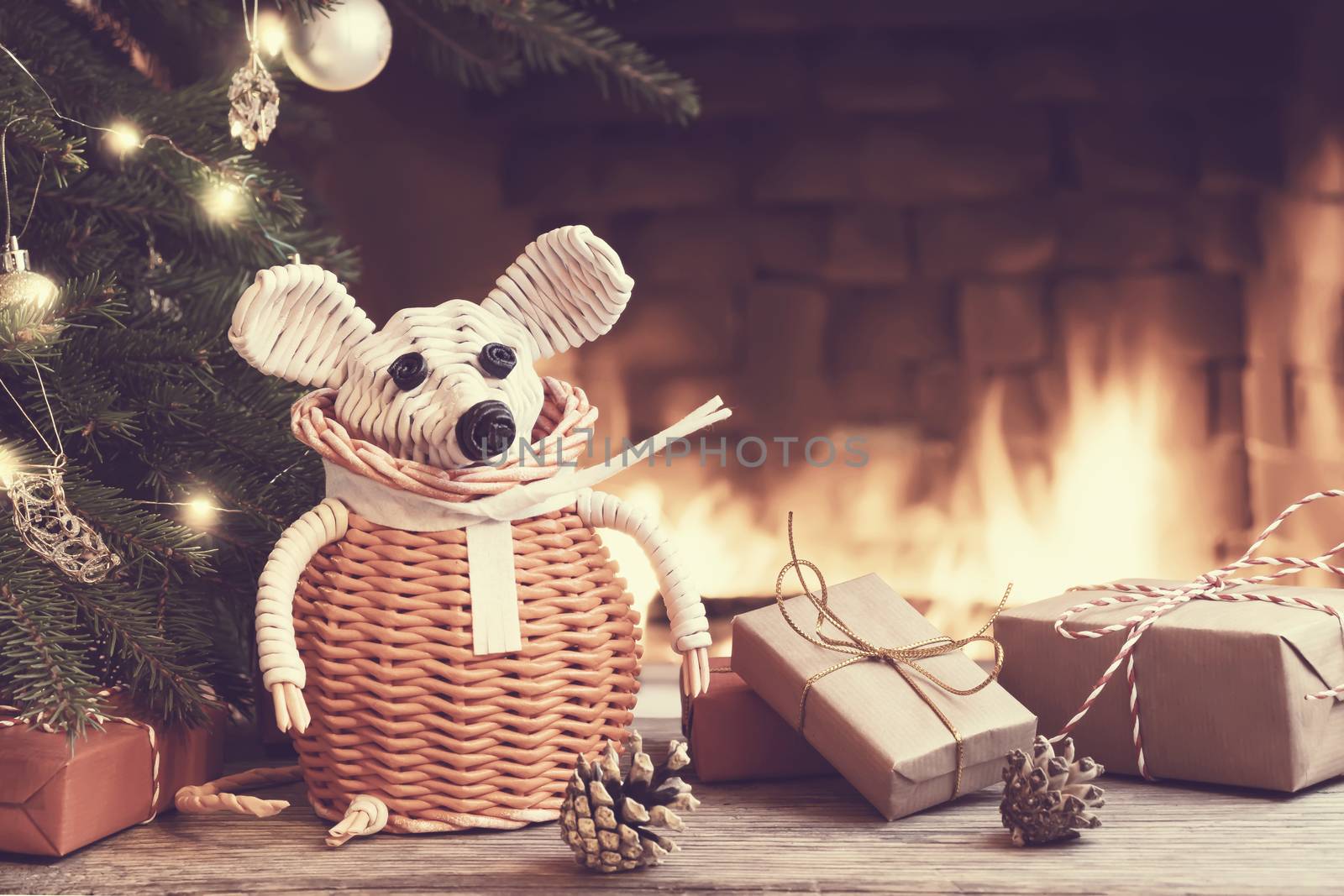 Christmas composition - a wicker white mouse - symbol of 2020 according to the Chinese horoscope next to gifts under the Christmas tree in room by fireplace.