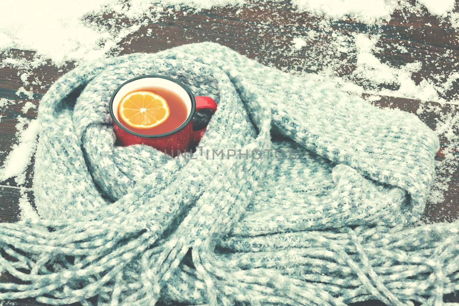 Red enameled cup of hot tea with lemon wrapped in a knitted scarf on a snowy wooden table.