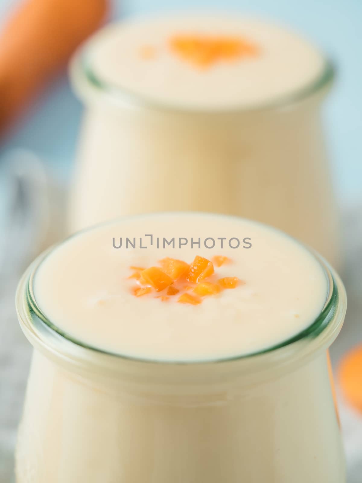 Yogurt with carrot. Vegetable yogurt. Two glass jar with yellow orange yoghurt or milkshake and fresh carrot on blue background. Copy space for text. Vertical.
