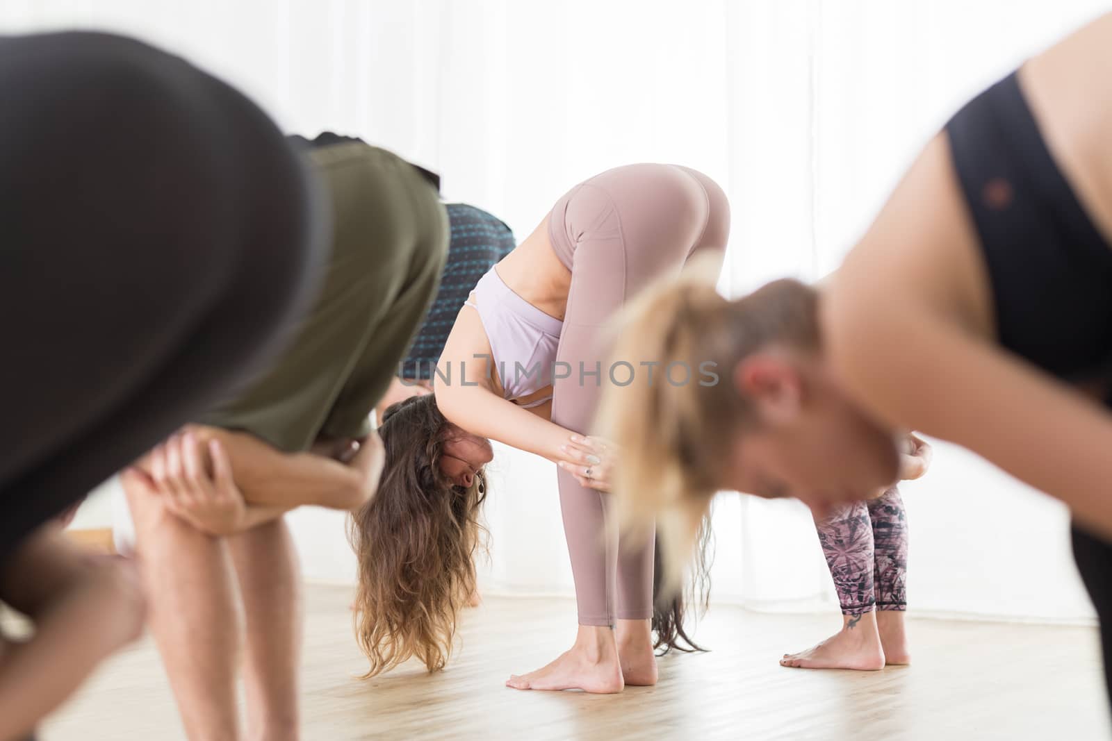 Group of young sporty attractive people in yoga studio, practicing yoga lesson with instructor, standing together in forward band leg streching pose. Healthy active lifestyle, working out in gym.