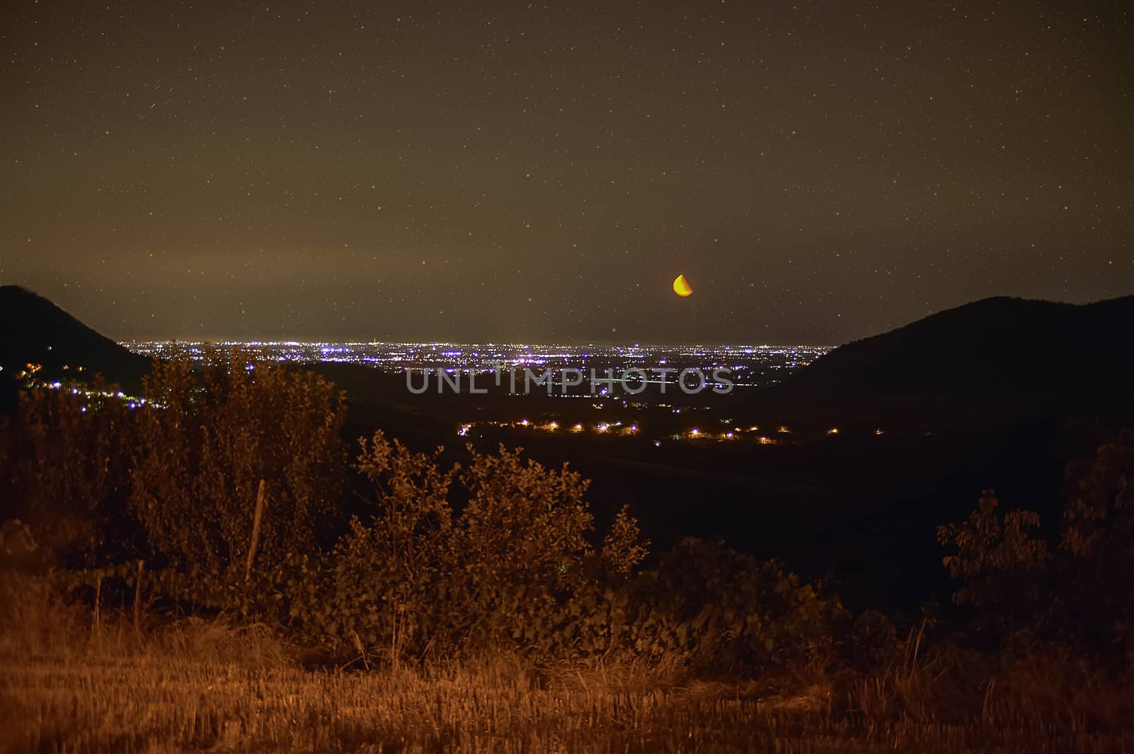Hilly landscape of northern Italy, Precisely of Este in Veneto. Very impressive night landscape where you can see the hillsides on the sides and the lights of the city that are lost in the horizon under a starry sky with warm colors and with the moon well visible.