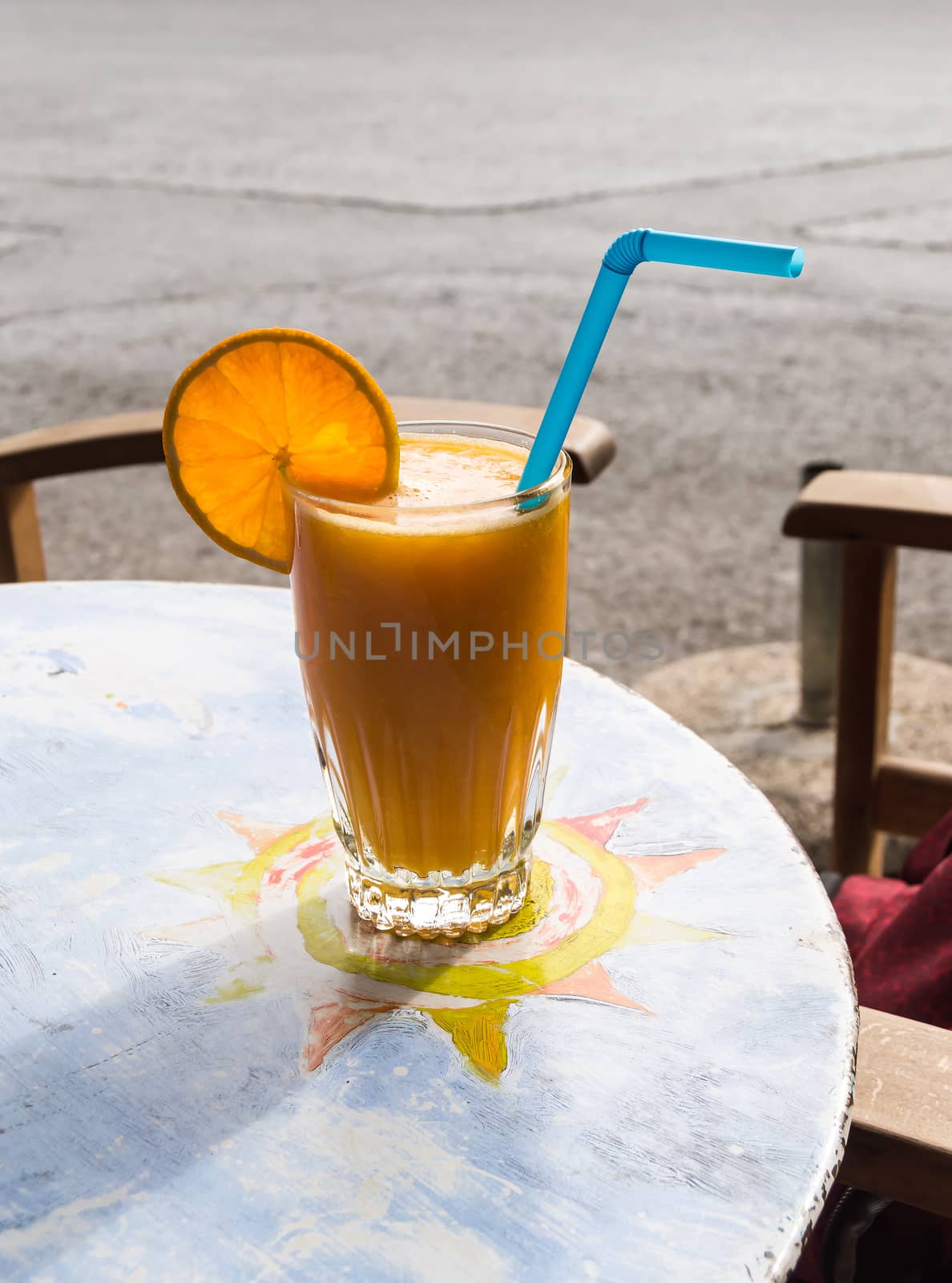 Orange juice smoothie drink with a slice of orange and a blue straw lay on a round table with the sun as a motive