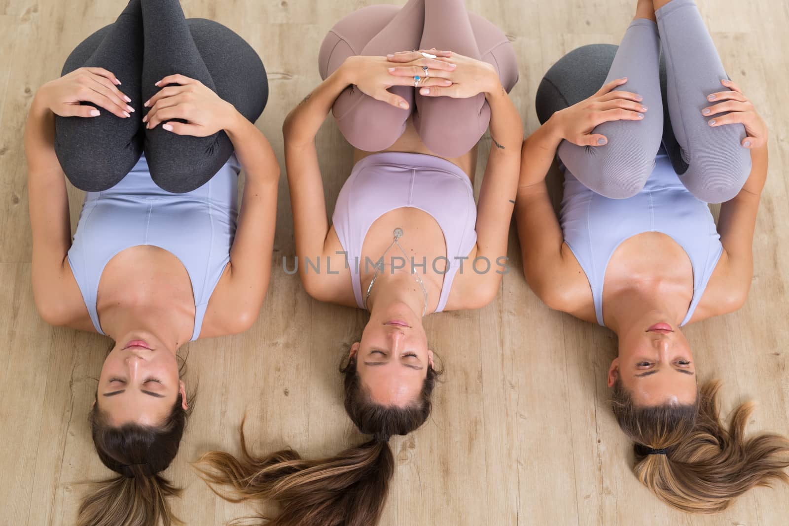 Group of three young sporty attractive women in yoga studio, lying on the floor, stretching and relaxing after the workout. Healthy active lifestyle, working out indoors in gym by kasto