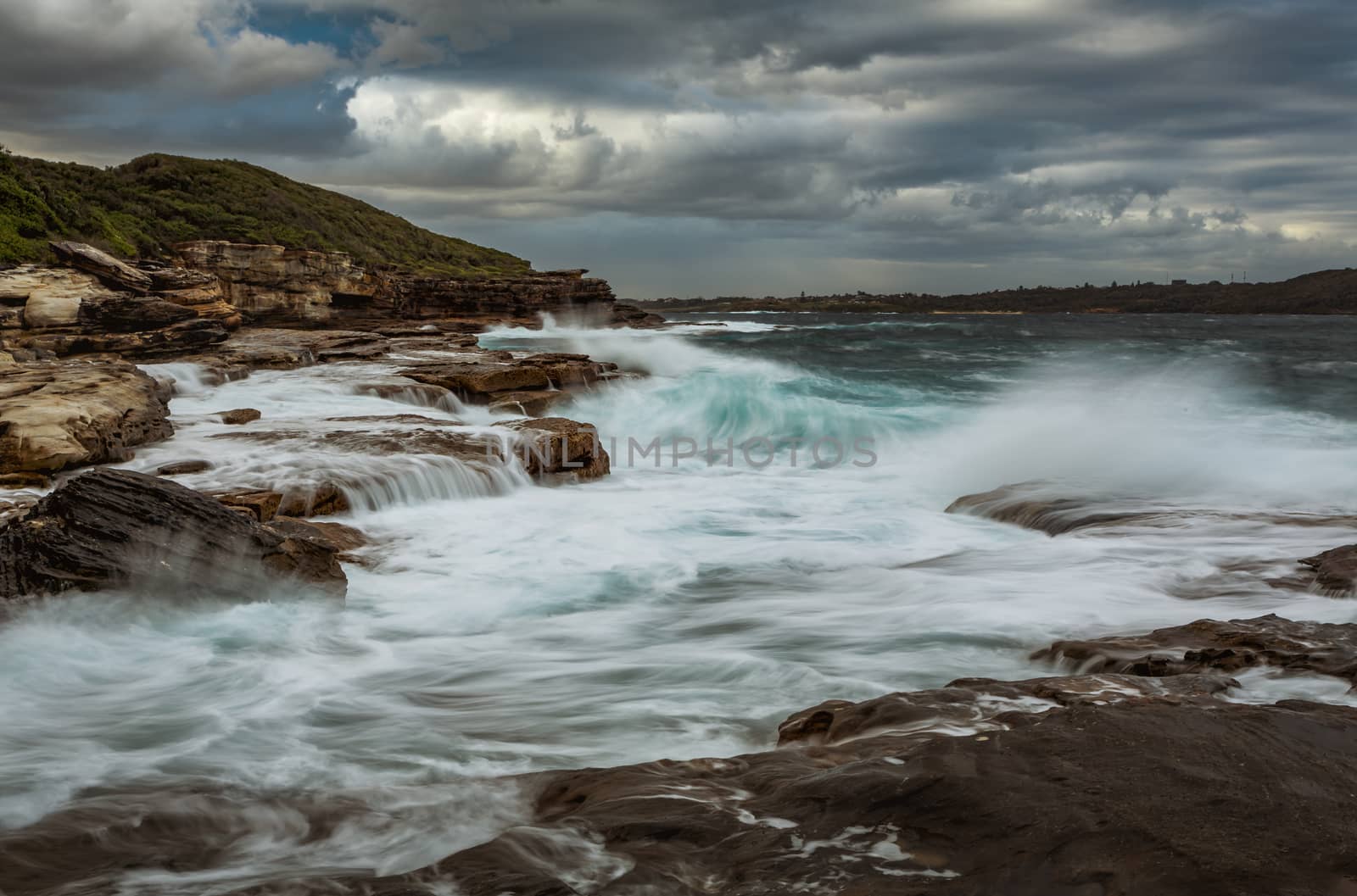 Turbulent waves wash haphazardly onto rocks and cascade back to the sea as fast flowing waterfalls.  Moody clouds loom overhead as rain falls in the distance