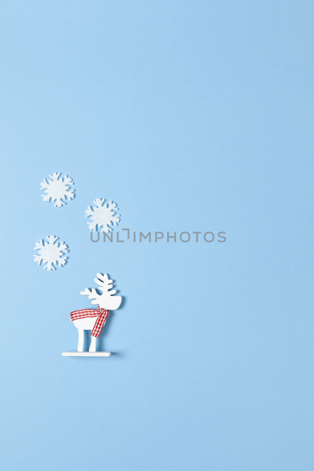 Christmas decoration, toy white deer in checkered scarf three little felt snowflakes on pastel blue background, copy space. Festive, New Year concept. Vertical, flat lay. Minimal style. Top view.