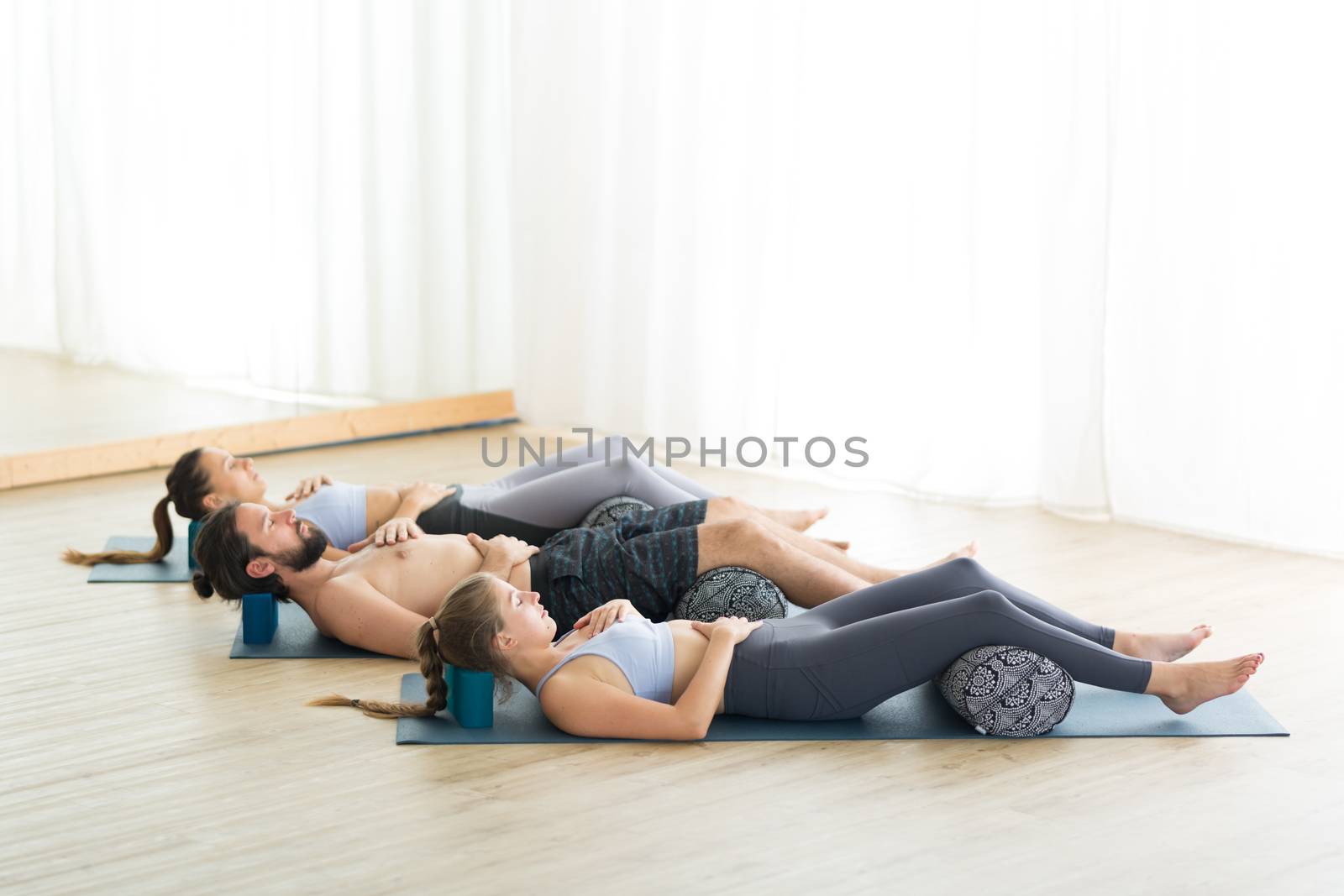 Restorative yoga with a bolster. Group of young sporty attractive people in yoga studio, lying on bolster cushion, stretching and relaxing during restorative yoga. Healthy active lifestyle by kasto
