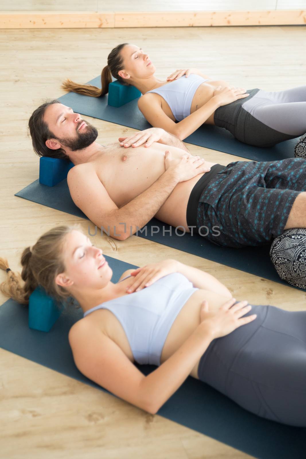 Restorative yoga with a bolster. Group of young sporty attractive people in yoga studio, lying on bolster cushion, stretching and relaxing during restorative yoga. Healthy active lifestyle.