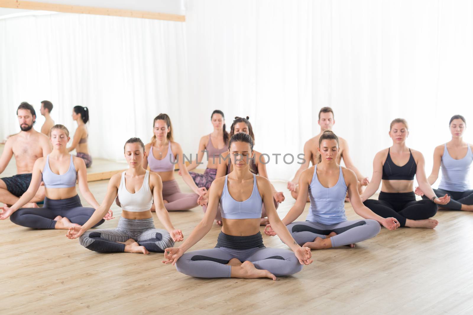 Group of young sporty attractive people in yoga studio, practicing yoga lesson with instructor, sitting on floor in Siddhasana, easy seated yoga pose. Healthy active lifestyle, working out in gym.