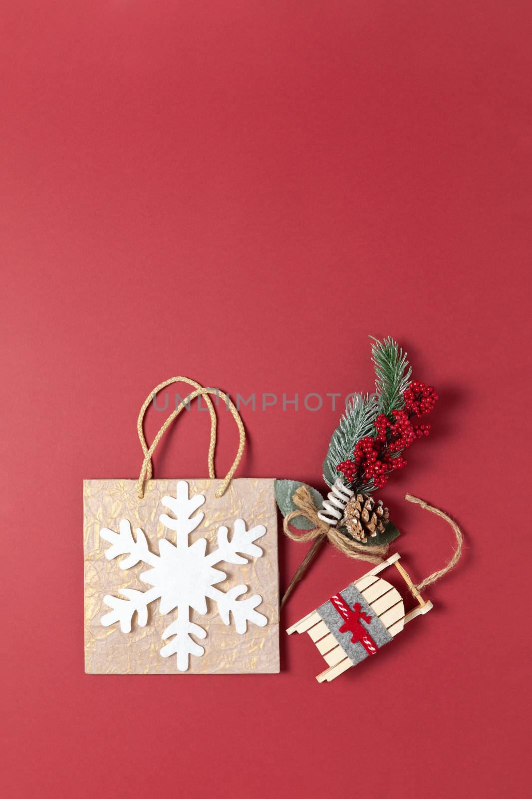 Christmas composition. Golden holiday package with white felt snowflake, bouquet with spruce twig and red berries, toy wooden sled on dark red background, copy space. Vertical, flat lay. Top view.