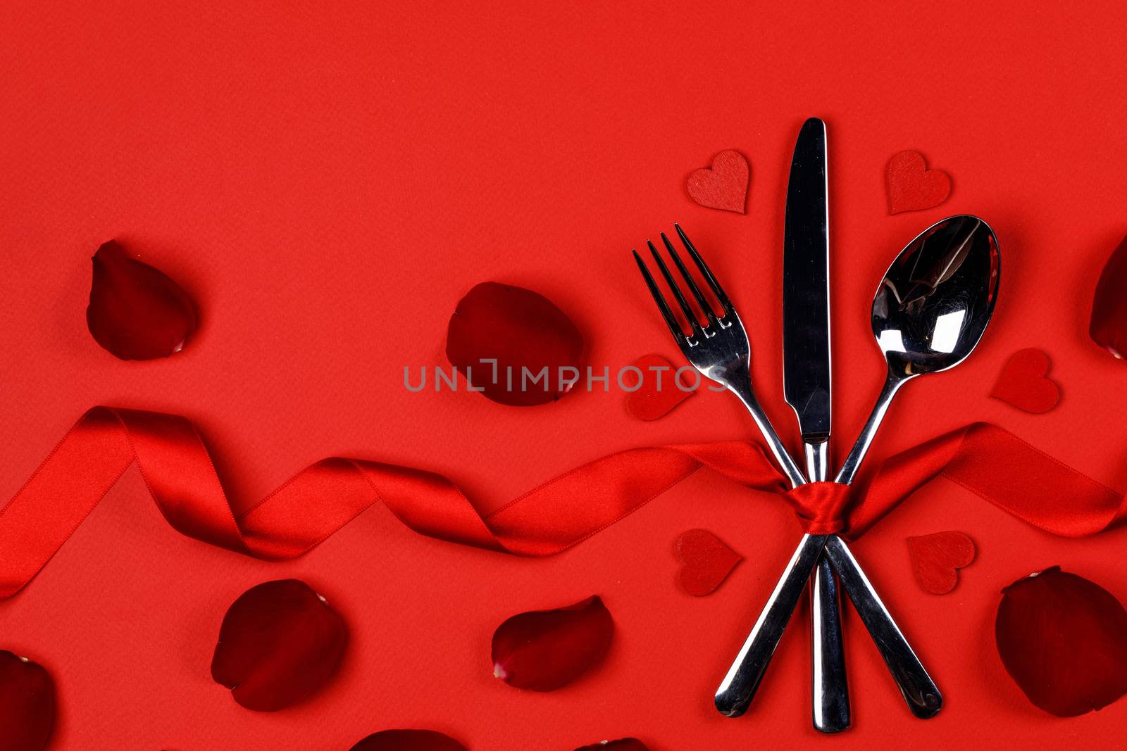 Cutlery set tied with silk ribbon rose flower petals and hearts on red background Valentine day dinner concept