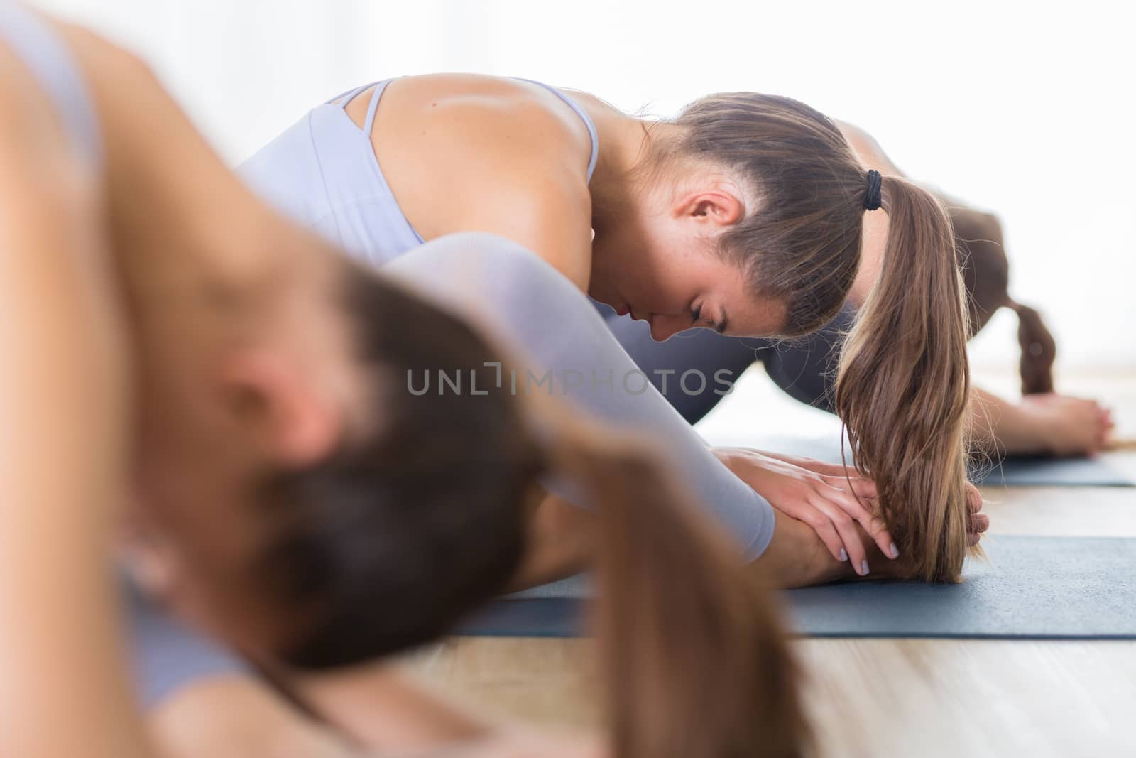 Group of young sporty attractive women in yoga studio, practicing yoga lesson with instructor, sitting on floor in forward bend yoga sana posture. Healthy active lifestyle, working out indoors in gym by kasto