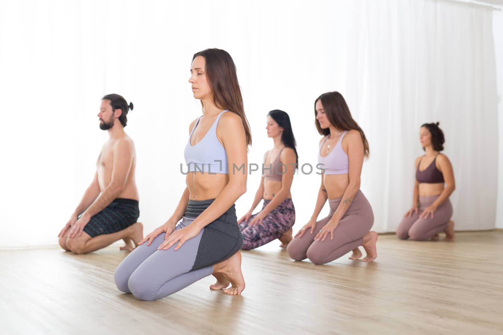 Group of young sporty attractive women in yoga studio, practicing yoga lesson with instructor, kneeling down, stretching and relaxing after workout . Healthy active lifestyle, working out in gym by kasto