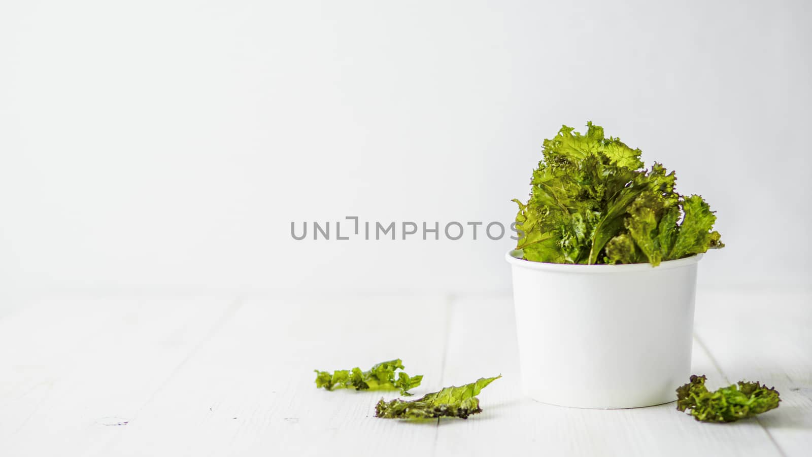 Kale Chips with salt in paper cup. Homemade healthy snack for low carb, keto, low calorie diet. White wooden background. Ready-to-eat kale chips, copy space for text. Banner